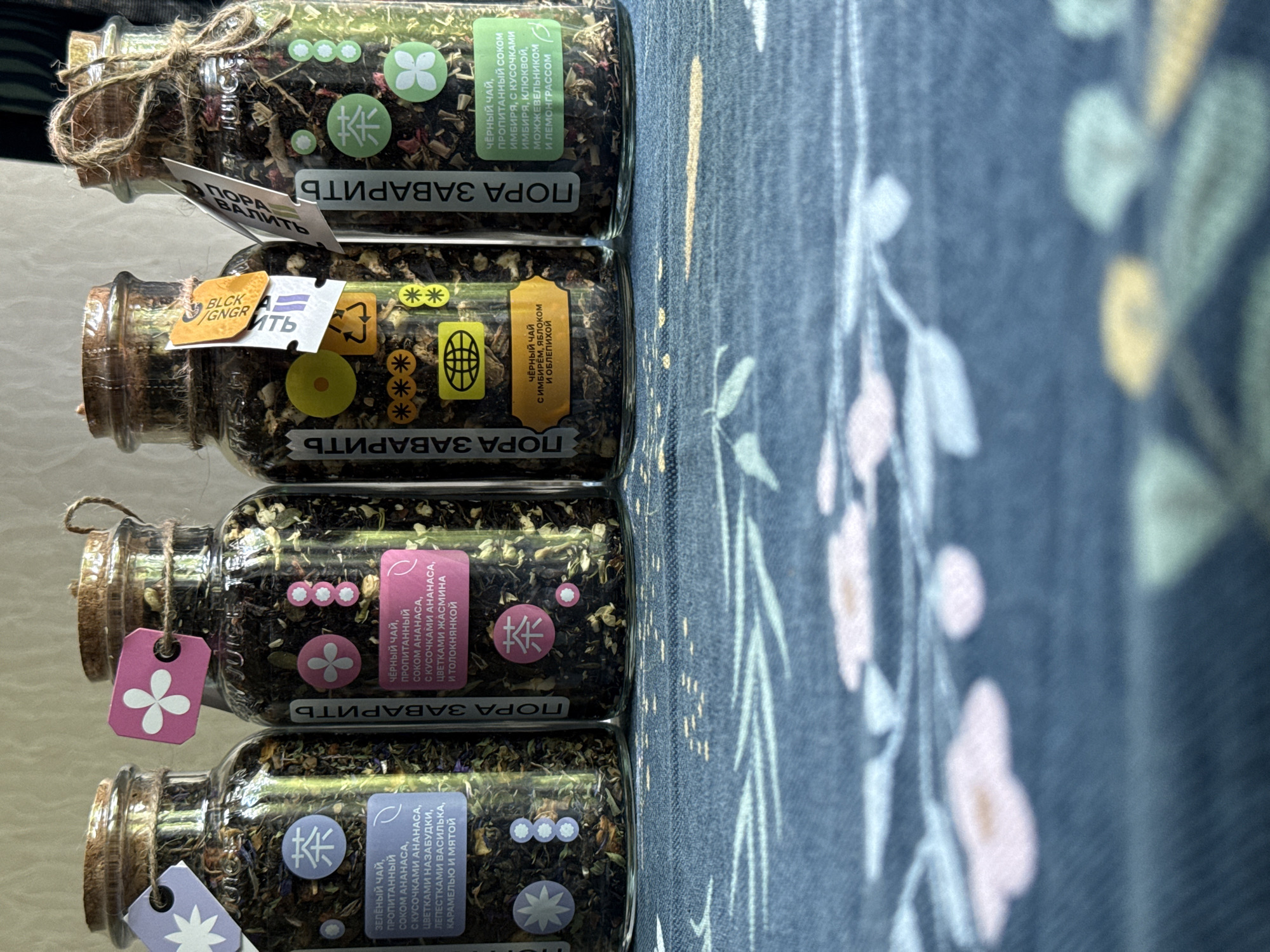 The image shows a stack of four glass jars, each filled with a dark mixture of herbs and spices, presumably tea. Each jar is labeled "ПОРА ЗАВАРИТЬ" (Time to Brew) and contains descriptions of the tea blend in Russian. The blends include black tea with ginger, apple, and sea buckthorn; black tea soaked in pineapple juice with jasmine flowers and bearberry; green tea soaked in pineapple juice with forget-me-nots, basil petals, caramel, and mint; and black tea with ginger juice, ginger pieces, cranberries, juniper, and lemongrass. The jars are tied with string and tags. The background is a floral-patterned surface, likely a tablecloth or bedspread. The location is likely in Russia, possibly Moscow, based on the Russian text on the labels.