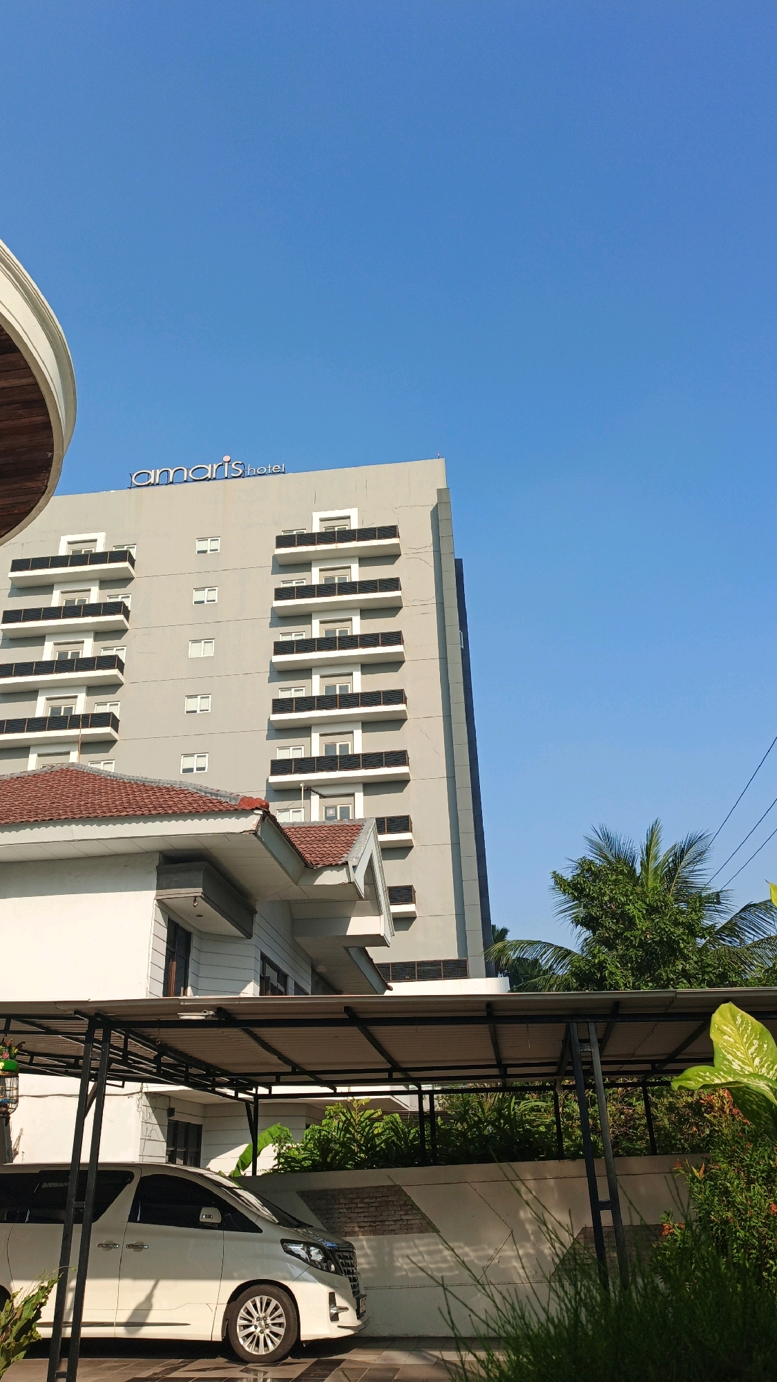 The photograph captures a clear, bright day with a vibrant blue sky dominating the upper portion of the frame. In the mid-ground, a tall, modern hotel building with a grey facade stands prominently. The hotel is identified by the blue sign "amaris hotel" at its apex. Multiple floors are visible, each featuring a series of balconies with dark railings and white trim, and small, uniformly placed windows.
In the foreground, a portion of a white modern building with a red-tiled roof is visible, positioned lower than the main hotel structure. To the left and slightly in front of this building, a white van, possibly an MPV, is parked under a covered carport. The carport has a dark metal frame and a light-colored roof. Lush green foliage and trees, including a palm tree, are scattered throughout the scene, suggesting a tropical or subtropical environment. The presence of power lines in the upper right corner indicates an urban or semi-urban setting. The overall impression is one of a well-maintained, contemporary hotel complex. Based on the distinct architecture and the presence of an "Amaris Hotel," this location is likely in Surabaya, Indonesia, a city known for such modern hotel establishments.