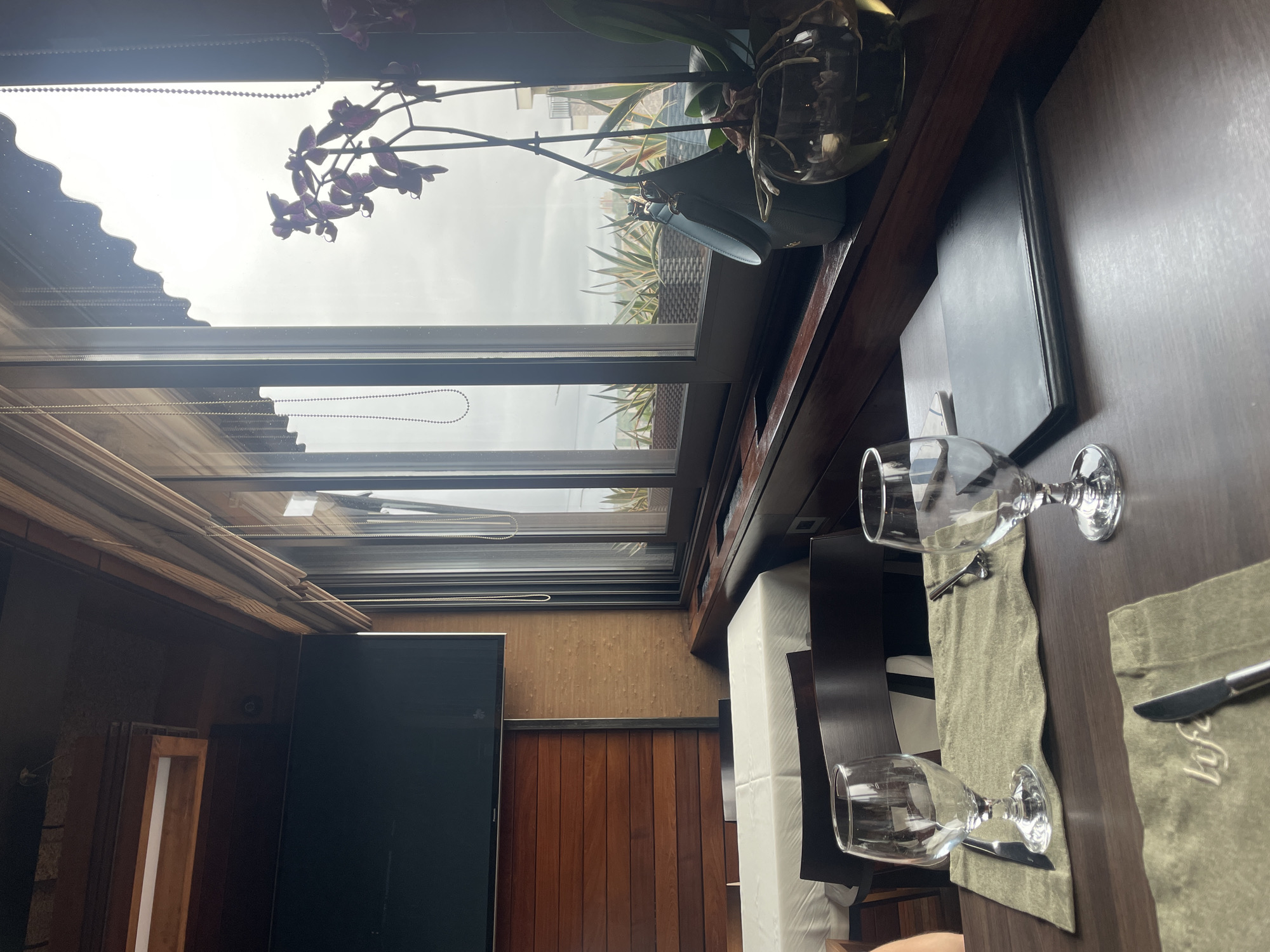 The image depicts a scene within a building, likely a restaurant or cafe, in Pleven, Bulgaria. The primary focus is a table setting with two wine glasses, knives, and placemats on a dark wooden surface. The room has wooden walls and a retractable glass roof. A potted orchid sits near the window, and a view of buildings and the sky can be seen through the glass.