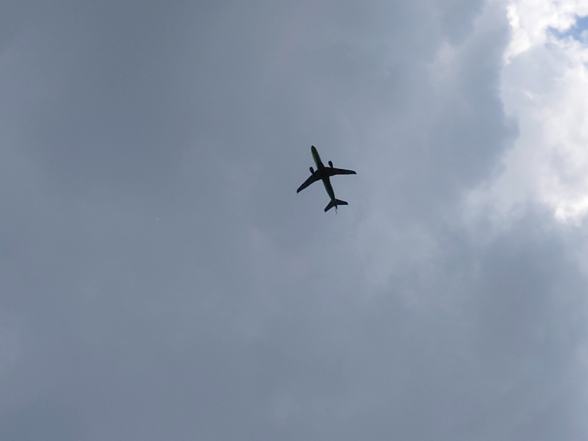 An airplane is silhouetted against a cloudy sky, viewed from below. The aircraft is a twin-engine jet with its wings extended and tail fins visible. It appears to be in mid-flight. The sky is overcast with varying shades of grey, suggesting an area of mixed cloud cover. The image does not contain any recognizable landmarks or features that would specifically indicate its location within Novosibirsk, Russia. The scene is natural and devoid of any human presence or interaction.