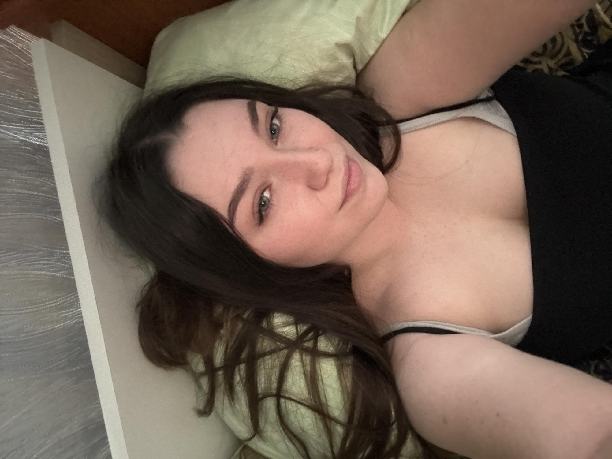 A young woman with long, dark hair is lying on a bed, looking at the camera. She is wearing a black tank top with a gray bra strap visible. The bed has a pale green pillow and a patterned blanket. The background appears to be a wall with a textured wallpaper. The woman's face is partially visible, and she has light-colored eyes and fair skin. Her hair is spread out on the pillow and the bed.