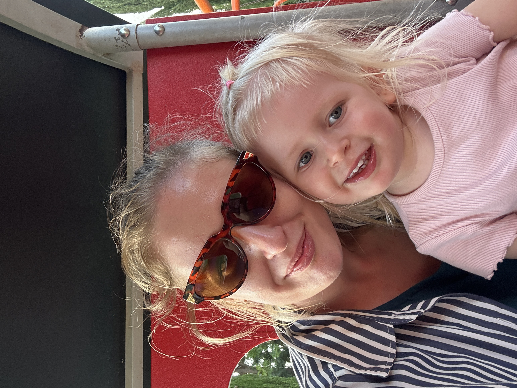A woman with sunglasses and a young child are posing closely together for a selfie. They are situated near playground equipment, with red and black panels and metal bars visible in the background. The woman wears a striped shirt. The child has blonde hair and is wearing a pink shirt. The photo appears to have been taken outdoors. While the playground equipment is common, it does not allow for pinpointing the location to Petah Tikva.