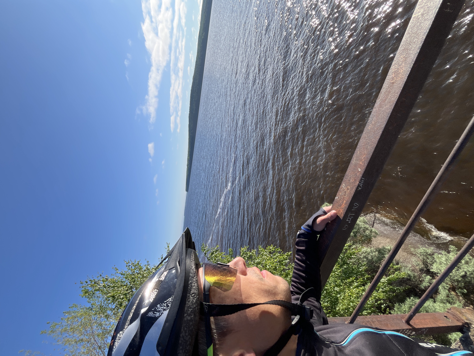 A man wearing a black and grey bicycle helmet and reflective orange cycling glasses is standing on a bridge, looking out over a large body of water. He is wearing a black cycling jacket with blue accents. The water is dark and choppy, with sunlight glinting off the surface. In the distance, a green, tree-covered shoreline is visible under a bright blue sky with scattered white clouds. The bridge railing, made of rusty metal, is in the foreground, and some graffiti is visible on it. Green foliage is growing on the embankment below the bridge. The location is on a bridge overlooking the Kama River in the city of Chaikovsky, Russia.