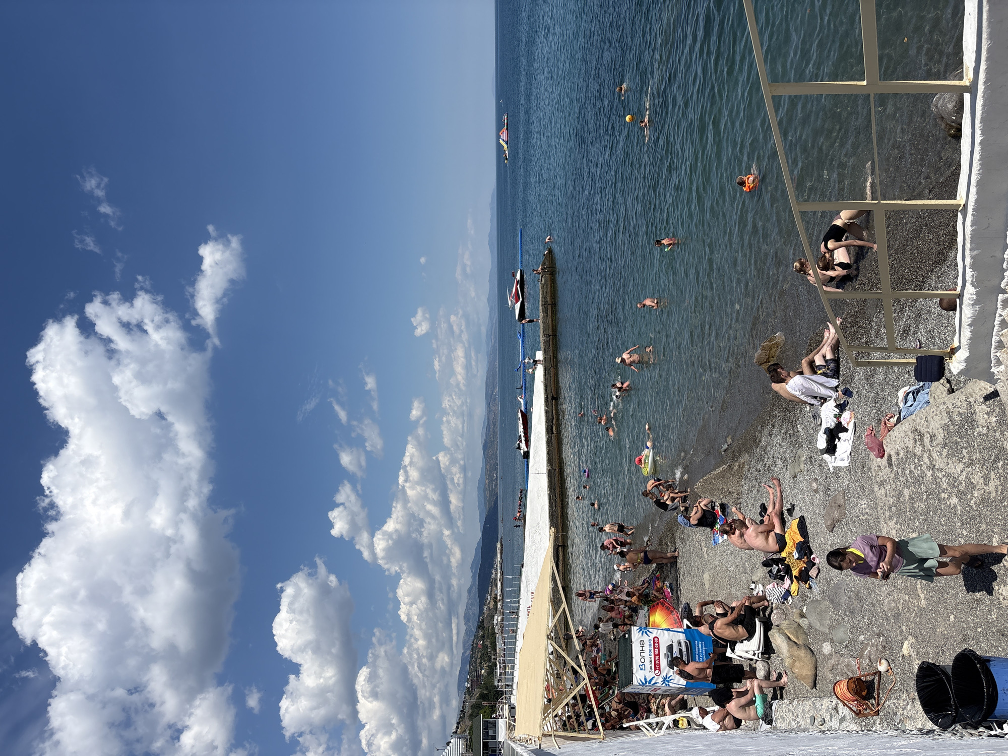 A crowded pebble beach on the Black Sea coast in Alushta, Ukraine. Numerous people are sunbathing on the shore and swimming in the clear blue water. Some are relaxing on towels, while others are engaged in various water activities, including playing with a yellow ball and floating on inflatable rings. In the distance, several boats, including a jet ski and a small passenger boat, are visible on the water. The coastline is lined with mountains, and the sky is a bright blue with fluffy white clouds. A white wall with a railing runs along the edge of the beach, separating it from the water in some areas. Beach umbrellas and shaded areas provide relief from the sun. The scene depicts a lively and typical summer day at a popular beach resort.
