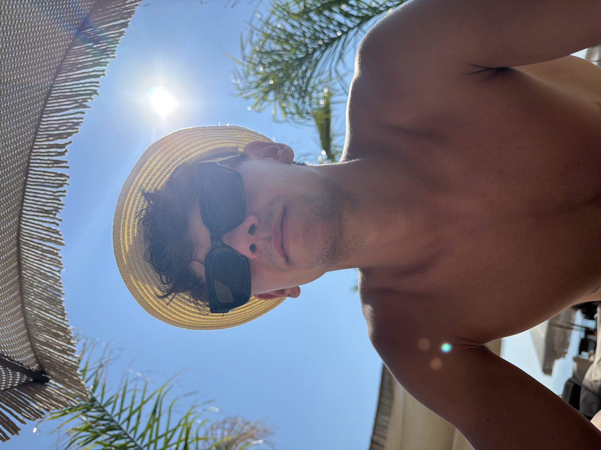A low-angle, waist-up, portrait-style selfie is captured outdoors on a sunny day at Ofrynio Beach, Greece. The subject, a shirtless young man with curly dark hair and stubble, wears a yellow straw hat and black rectangular sunglasses. He is positioned centrally in the frame, looking directly at the camera. Above and behind him, the bright sun is visible, creating lens flares and glares. A straw beach umbrella with fringes hangs in the upper part of the frame, and palm fronds are visible on either side, framing the scene. The background consists of a clear blue sky. Out of focus in the lower background, lounge chairs and umbrellas suggest a beach resort setting. The lighting is bright and direct, indicative of midday sun.