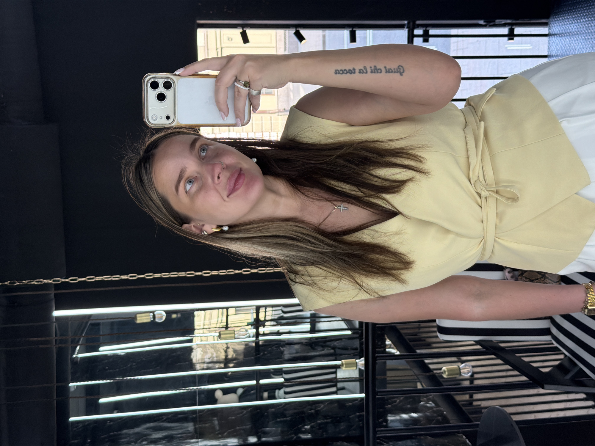 The image is a selfie taken by a woman indoors. She is holding a smartphone with three camera lenses. The woman has fair skin, light brown hair with blonde highlights, and blue eyes. She is wearing a yellow wrap-style top and a white skirt or pants. She has a tattoo on her forearm. She is wearing a necklace with a cross pendant and earrings. There are rings on her fingers and a gold watch on her wrist.
The background shows a reflective surface with black and white stripes, and a window with a view of a building outside. The scene is lit by artificial lighting. The city and specific location are unknown.