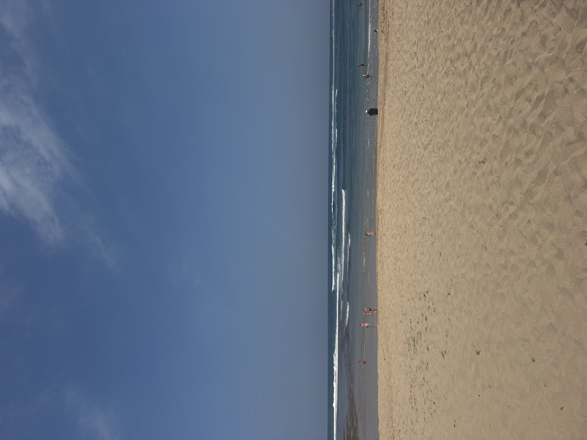 The image captures a beach scene in Kuta Selatan, Indonesia, under a clear blue sky with scattered wispy clouds. The foreground is dominated by a wide expanse of golden sand, showing textured ripples from wind and tide. In the mid-ground, a gentle shoreline meets the ocean, with several lines of white-capped waves rolling towards the beach. A few people are visible on the sand and in the shallow water. On the left side, near the water's edge, a red flag is planted in the sand. Further to the right, a group of three individuals stand in the shallow water, and another person is wading further out. Closer to the right edge of the frame, a solitary figure sits facing the ocean, their back to the camera. On the far right, another individual is visible in the water. The ocean appears calm beyond the breaking waves, with the horizon line clearly defined against the sky. The overall impression is one of a peaceful day at a tropical beach.