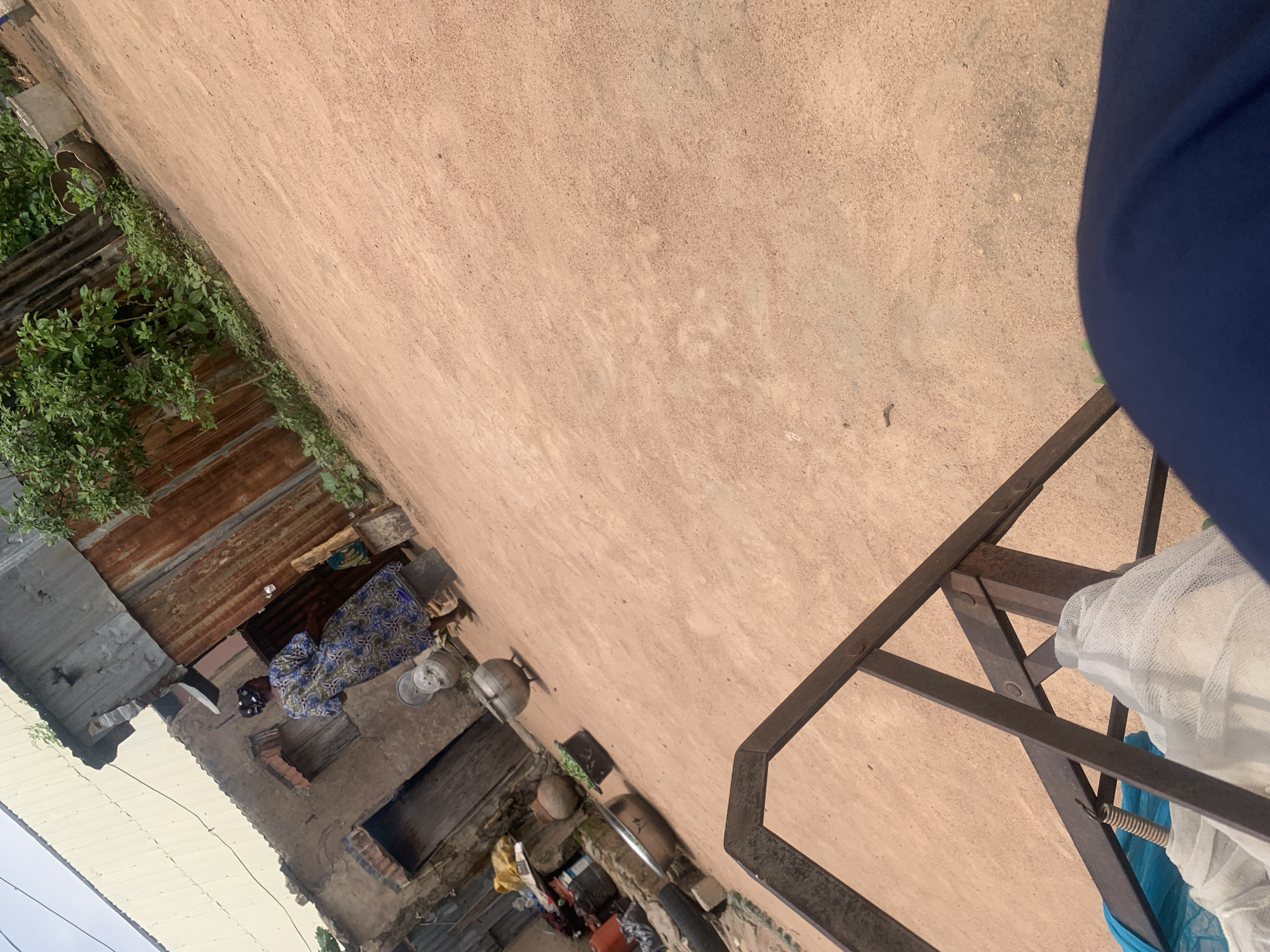 The image depicts an outdoor scene in Jalingo, Nigeria. In the mid-ground, a person is seated in front of a rustic structure with a corrugated metal roof, wearing a patterned blue and white dress. Several metal pots and cooking utensils are scattered around the area. The surrounding environment appears to be a courtyard or an outdoor living space, characterized by rough, sandy ground and a pinkish-tan wall. To the left, there's more corrugated metal roofing and green foliage. In the foreground, a dark metal railing with some white mesh material draped over it is visible. The lighting suggests it is daytime, possibly late afternoon due to the warm tones of the wall. The overall impression is of a simple, everyday setting in a rural or semi-urban environment.