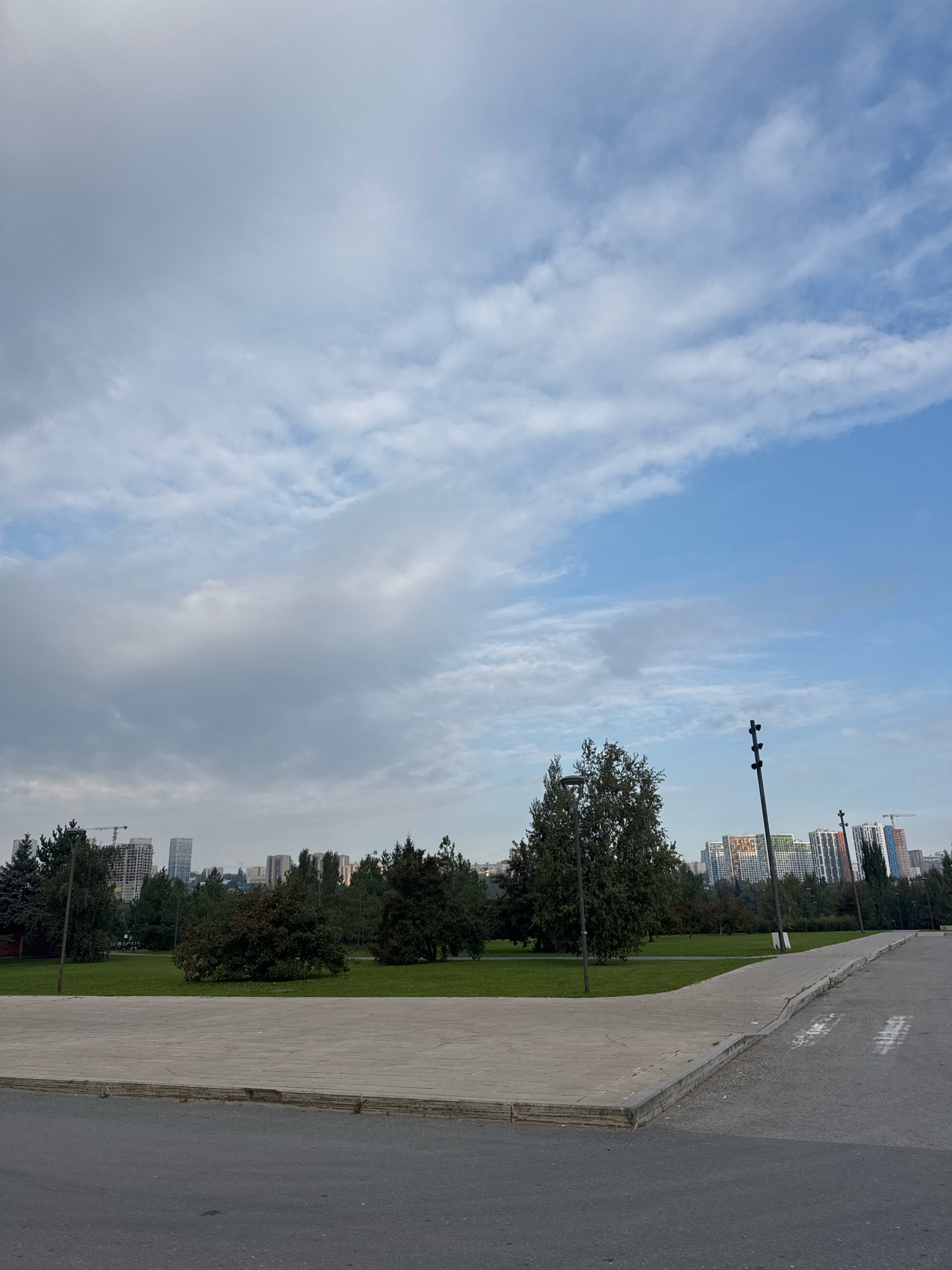 The image shows a street scene in Ufa, Russia, likely taken from a sidewalk overlooking a park area. In the foreground, there is an asphalt road with a concrete sidewalk bordering a grassy park. The park features several trees and a well-maintained lawn. In the background, a cityscape is visible with apartment buildings of various heights and colors. The sky is partly cloudy, with a mix of gray and blue hues. There are no people or animals visible in the scene. The overall atmosphere appears to be calm and urban.