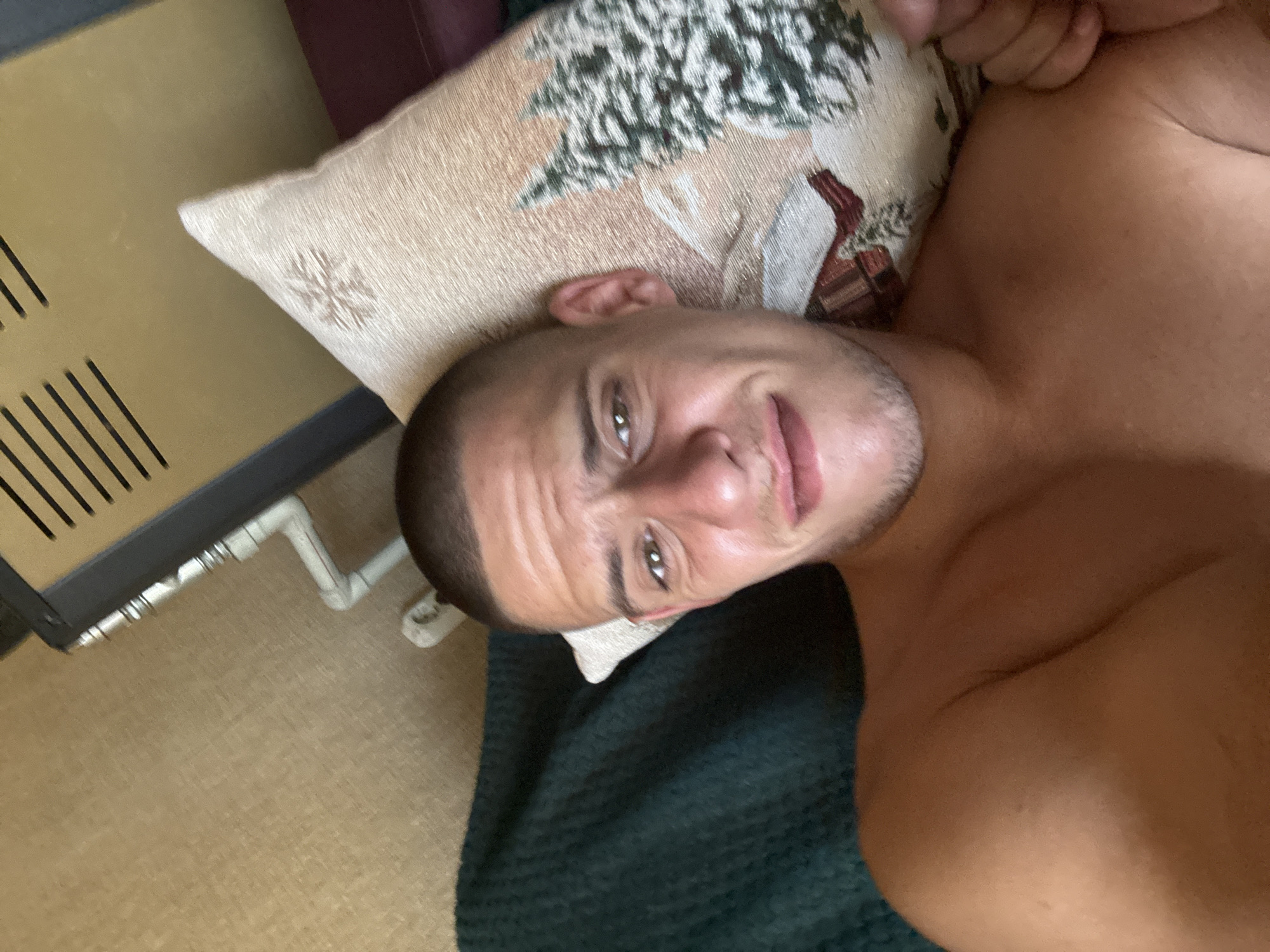 The image shows a close-up, high-angle, selfie-style shot of a man lying down. He has short, dark hair and is looking towards the camera with a slight smile. His chest and shoulder are visible. He is resting his head on a pillow with a festive design featuring trees and a snowflake. In the background, a dark green textured fabric is visible behind the pillow, and to the right, a beige, textured surface with a metallic grate and white piping is present. The lighting suggests an indoor setting. The image does not contain any information that would allow for the determination of its location in Sofia, Bulgaria.