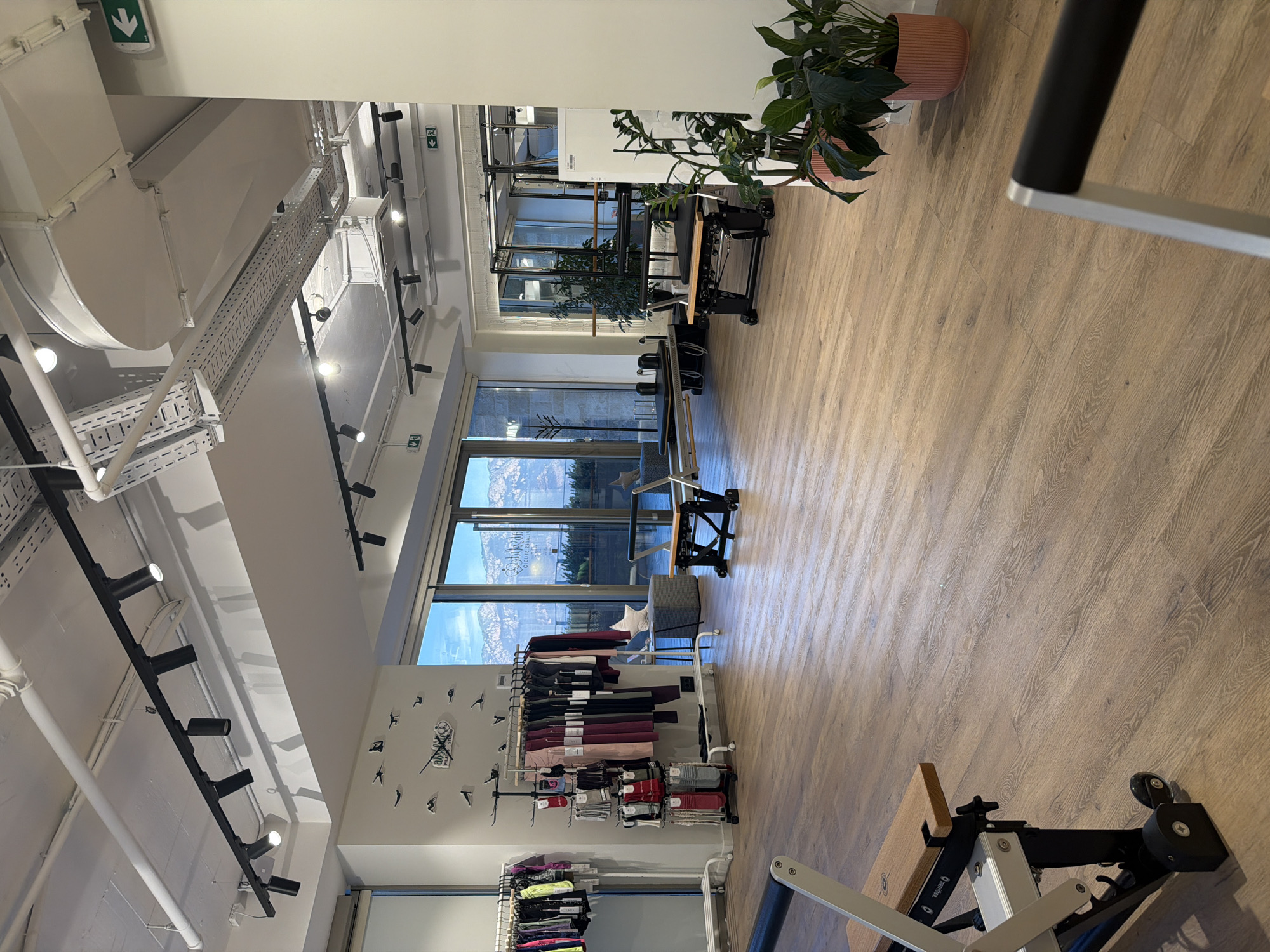 The image depicts the interior of a modern fitness studio, possibly a Pilates studio, in Boreti, Montenegro. The space features a light wood-toned floor and is equipped with various exercise machines, including reformers and apparatus common in Pilates training. Large windows offer a view of mountainous terrain and a bright sky. Racks of athletic apparel are visible along one wall, suggesting retail or display space. The ceiling is high, with exposed ductwork and track lighting. Several potted plants add a touch of greenery to the environment. The scene appears to be unoccupied at the time the photograph was taken.