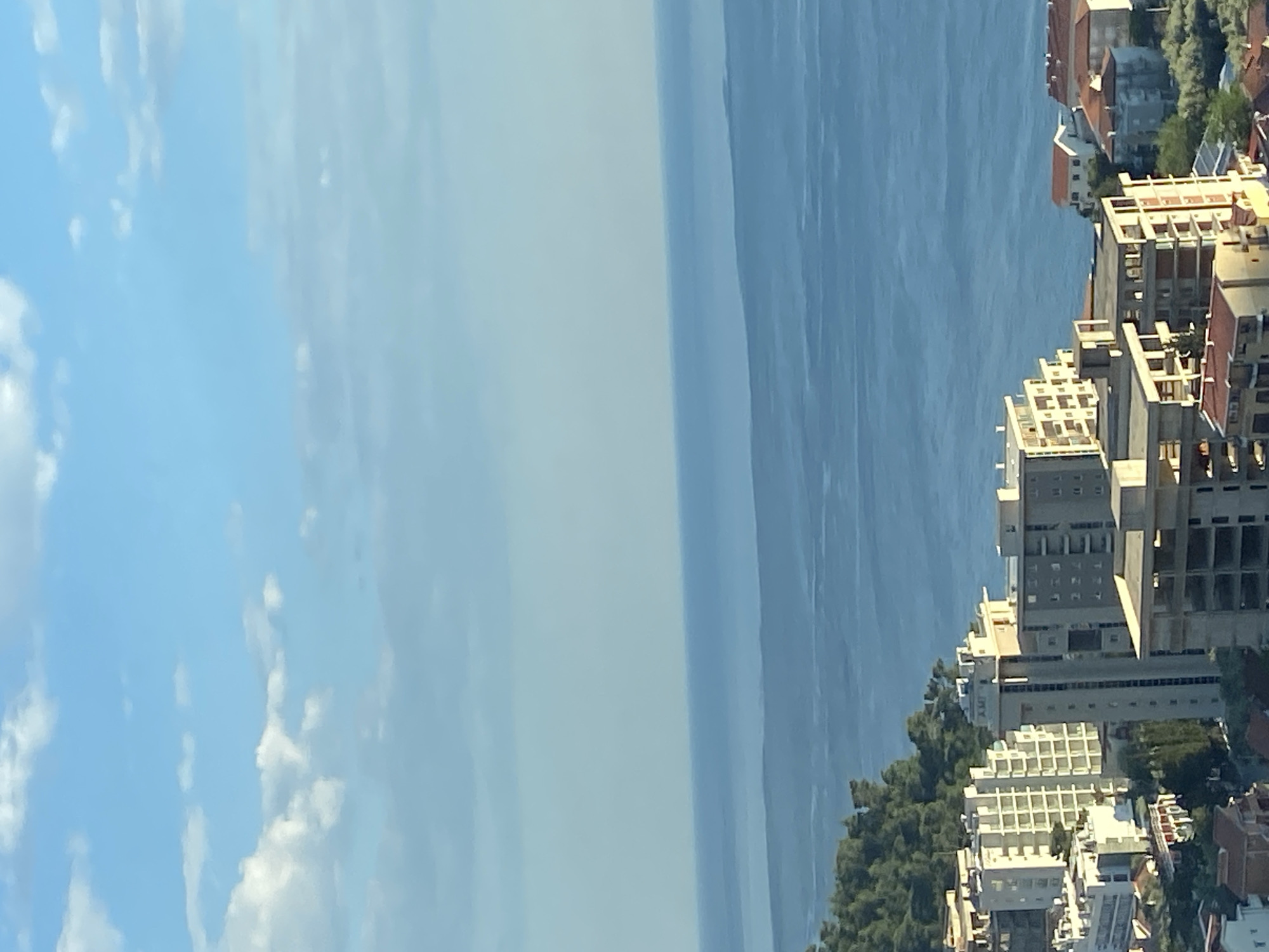 The image captures a high-angle, rotated view of Petrovac na Moru, Montenegro. The scene shows a cityscape dominated by light-colored buildings with red-tiled roofs, modern high-rise structures, and green trees. The Adriatic Sea stretches out beyond the city, merging into a hazy sky in the distance. The sky on the left side of the image is a lighter blue with scattered clouds.