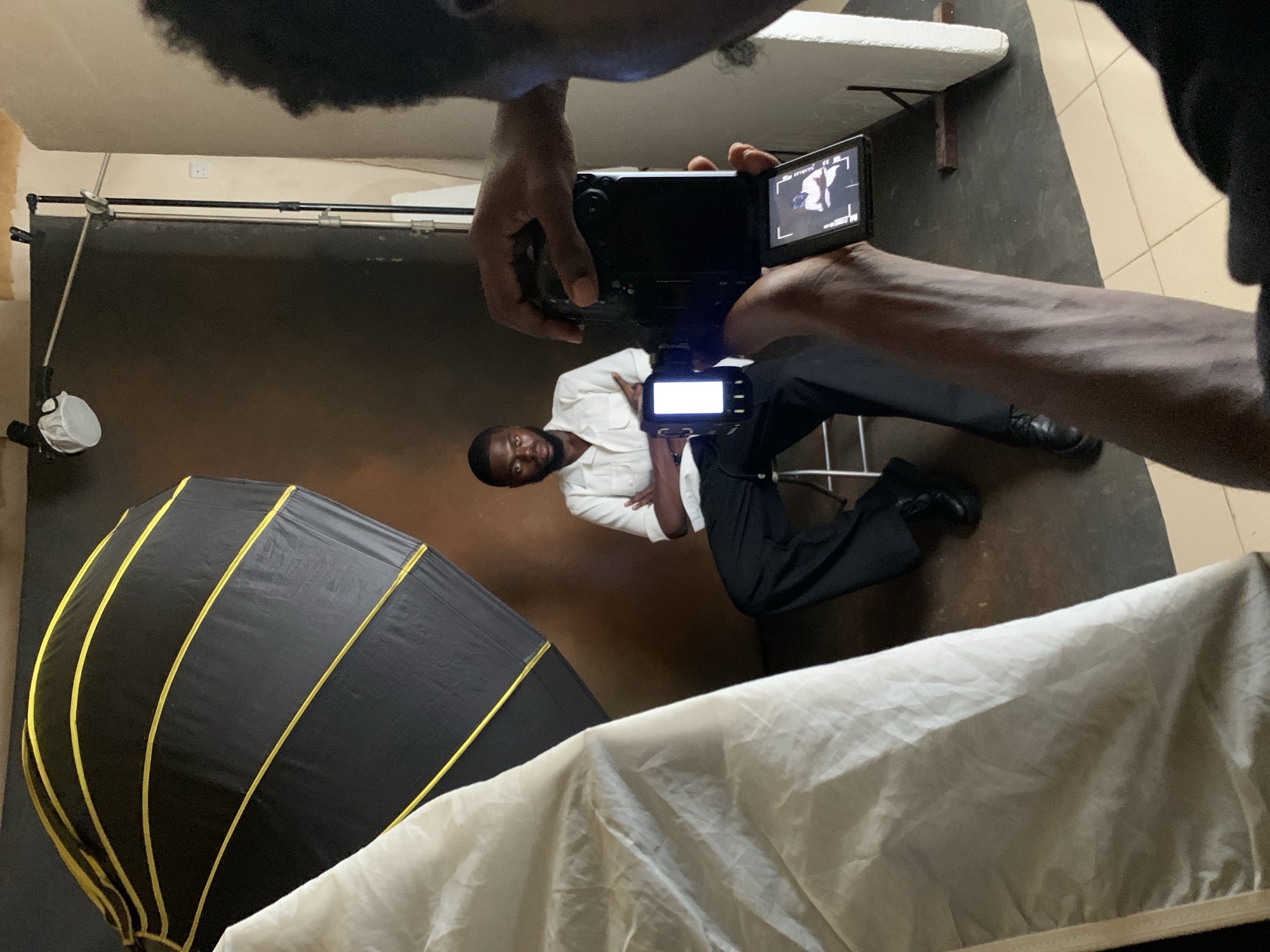 In what appears to be a studio setup in Kwang, Nigeria, a man is seated on a stool in front of a dark brown backdrop. He is wearing a white collared shirt and black trousers, with his arms crossed. To the left, a large, black, octagonal softbox with yellow trim is positioned, partially obscured by a white fabric diffusion screen. In the foreground, the back of a person is visible, holding a black video camera and framing the seated man. The camera's LCD screen displays a preview of the shot, showing the seated man. The environment features tiled flooring and a white foam board or reflector against the right wall.