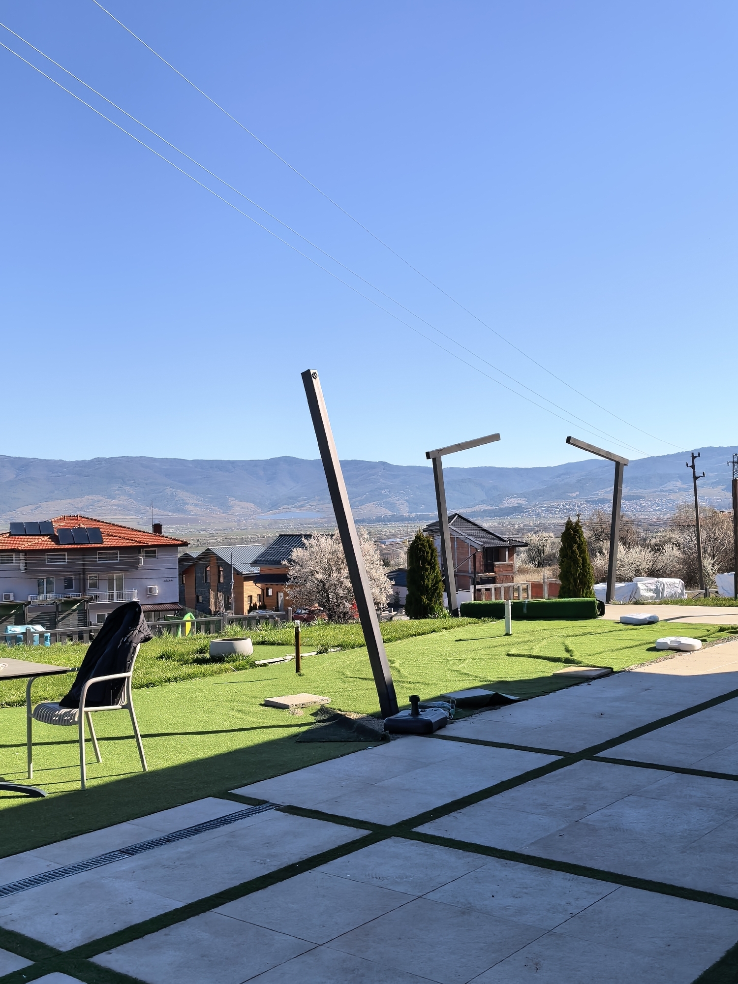 Here's a detailed description of the image:
**Location:** The image appears to be taken outdoors in an elevated residential area overlooking a city. The exact city is unknown.
**Environment:** The scene includes a well-maintained outdoor space with a combination of artificial turf and tiled paving. The weather is clear and sunny. In the background are mountains and a city view.
**Objects:**
* Wooden support structures (likely parts of an incomplete structure) stand upright on the turf.
* Rolls of artificial turf are visible.
* A table and chair set with a jacket are present.
* Houses with solar panels are in the midground.
* Power lines run across the sky.
**Subjects:** There are no people visible in the frame.
**Actions/Interactions:** There is no visible activity taking place in the scene. The image appears to depict a construction or landscaping project in progress.
