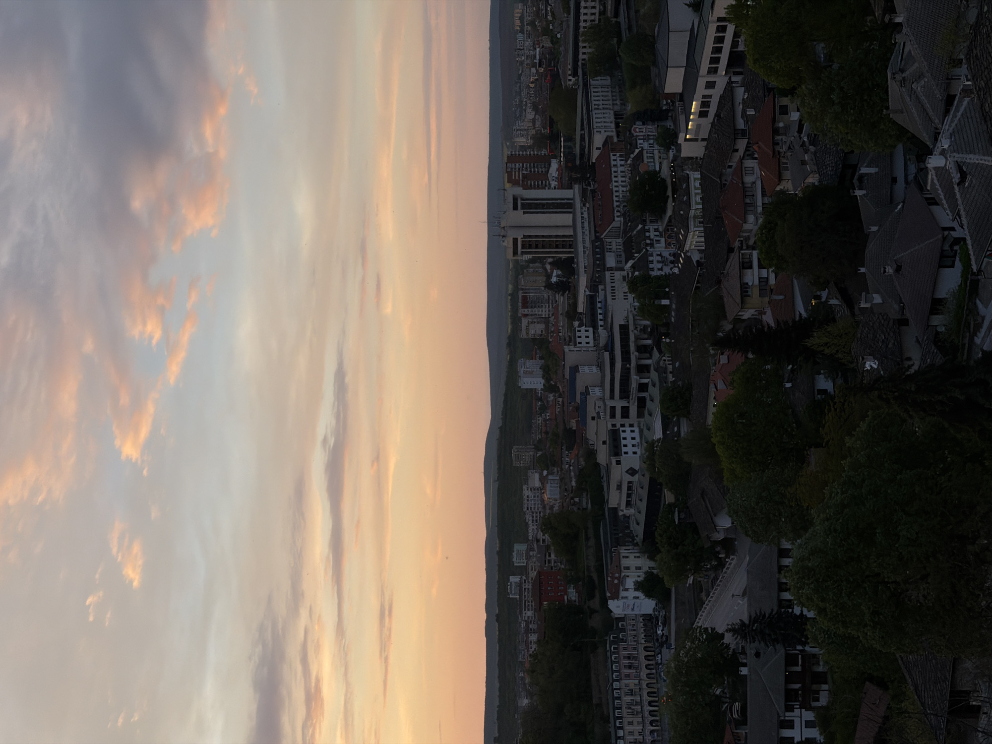 The image shows an aerial view of Lovech, Bulgaria at sunset. The sky, occupying the left half of the frame, features a mix of clouds tinged with pink and orange hues from the setting sun. A dark line of hills or mountains separates the sky from the city below. On the right half of the frame is a dense urban area with a mix of residential buildings, trees, and streets. Building rooftops vary in color, with some appearing gray and others reddish. The overall lighting suggests it's late evening or dusk. No people or specific actions are readily discernible from this high-angle perspective.
