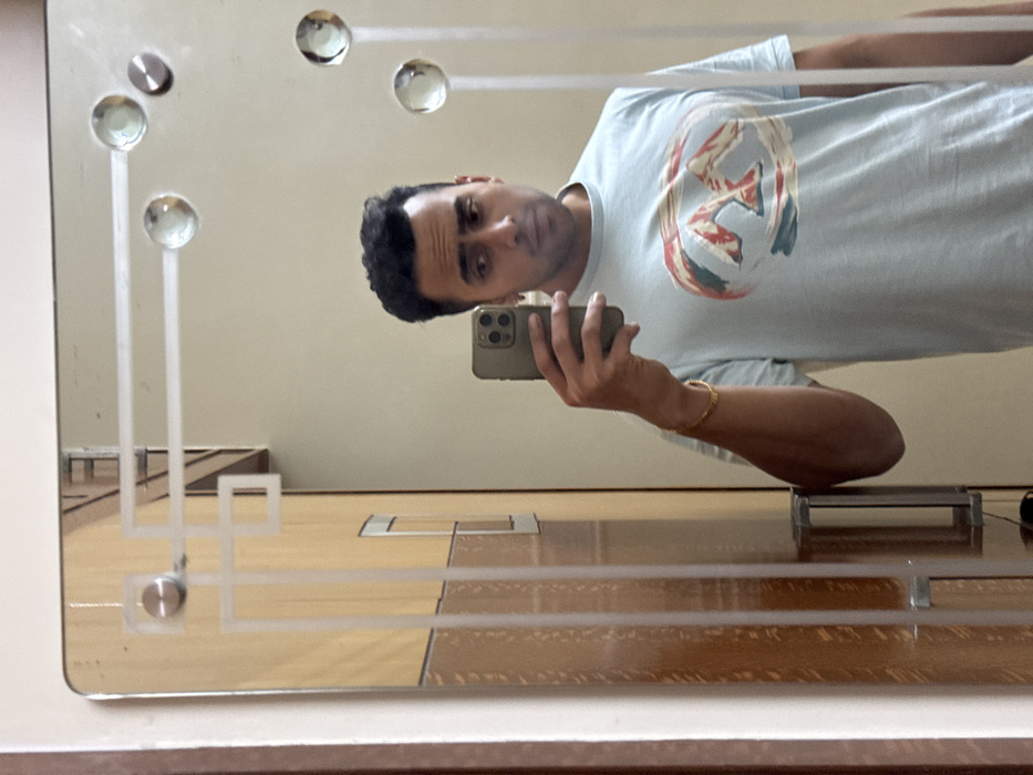 A selfie reflection shows a man with dark hair, holding a smartphone. He wears a light blue t-shirt featuring a graphic design. The mirror has decorative round glass orbs and linear etched patterns. The background reveals a room with a wooden floor, likely a basketball court based on the visible lines. A grey object rests on the floor, possibly a piece of equipment.