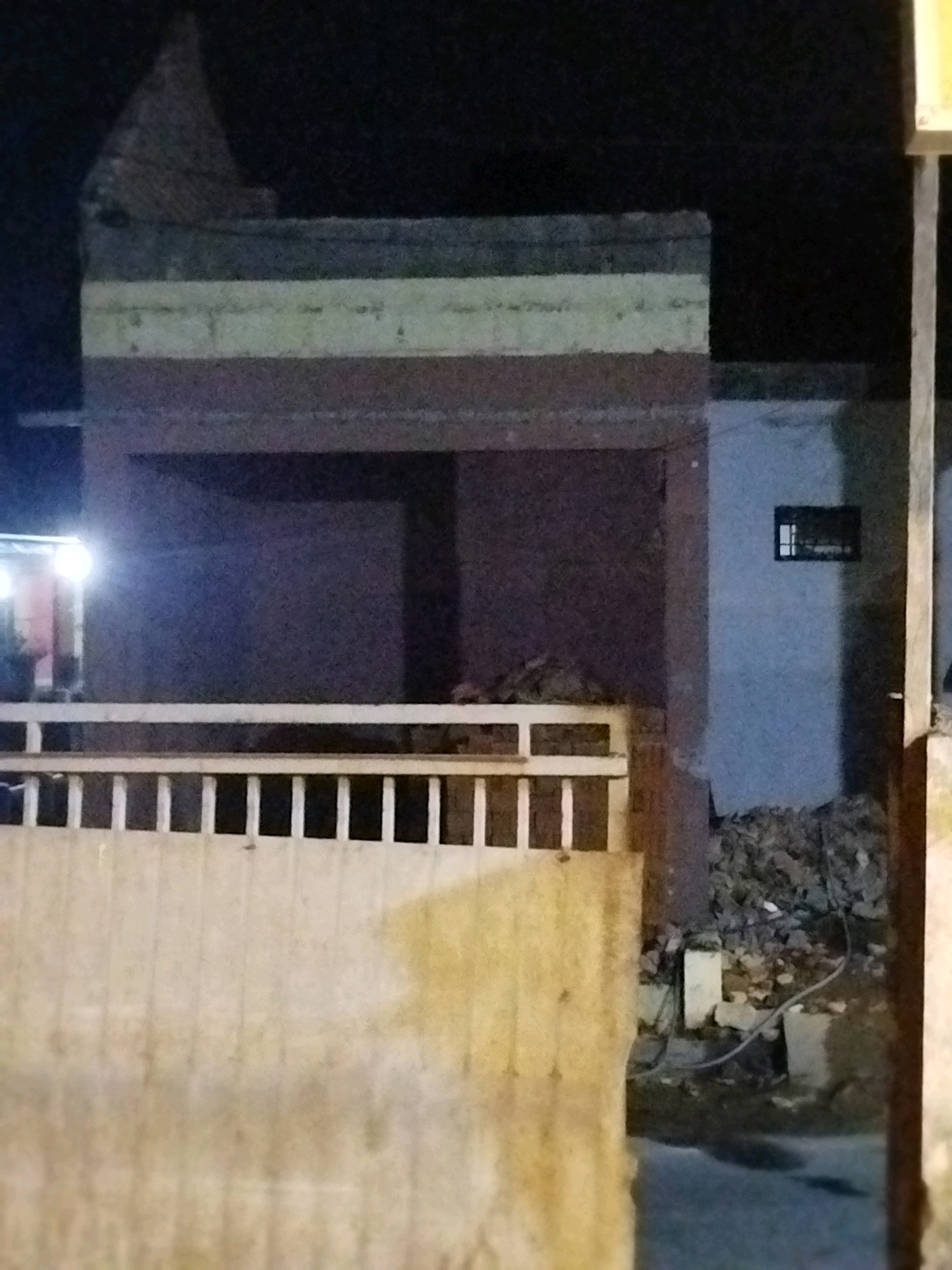 The image depicts a dark, nighttime scene in Mojopanggung, Indonesia, likely near a construction or demolition site. In the foreground, a light-colored, textured wall or barrier occupies the bottom half of the frame, with vertical lines suggesting a gate or fence. Above this, a white railing with vertical supports extends horizontally across the middle ground.
The background reveals a two-story structure, partially illuminated. The upper level features a prominent horizontal band, composed of alternating dark and light-colored sections, possibly indicating different materials or paint schemes. Above this band, a peaked, dark structure, possibly a roof or a decorative element, is visible against the dark night sky.
The lower level of the building shows an open, dark space, likely a covered area or porch, with a small, illuminated section to the left, possibly an entrance with a warm light. To the right of the building, a light blue wall is visible, with a small, dark rectangular window near the top. The ground area to the right is cluttered with debris, including rocks, rubble, and what appears to be wiring, suggesting ongoing construction or demolition work. The overall impression is one of an unfinished or dilapidated building at night.