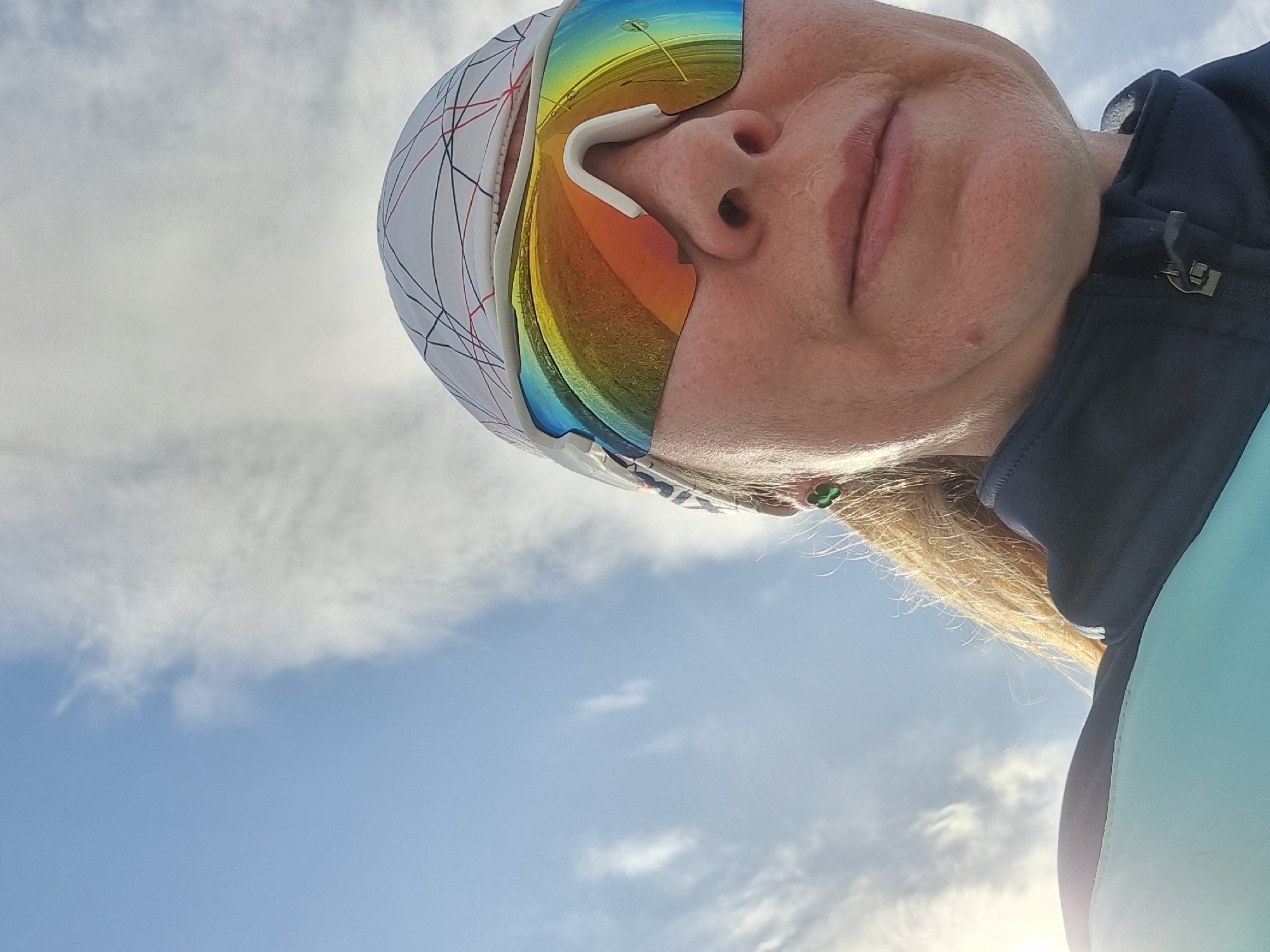 The image, captured in Chaikovsky, Russia, features a low-angle, close-up perspective of an individual's head and upper torso against a sky background. The subject has blonde hair and wears a white head-covering with a geometric pattern of black and red lines. Large, wrap-around sports sunglasses with highly reflective, rainbow-mirrored lenses obscure the eyes. The reflection in the lenses depicts a curved road or path flanked by green vegetation, illuminated under a sky. Visible skin on the face and neck shows texture, including some facial lines and fine hair on the chin. A small, green, clover-shaped earring is present on the visible earlobe. The subject wears a dark blue zip-up jacket with a high collar and a lighter teal-colored panel below. The background consists of a partially cloudy blue sky with scattered white, wispy clouds. A bright light source, consistent with the sun, is visible in the lower-left portion of the frame, causing a lens flare.