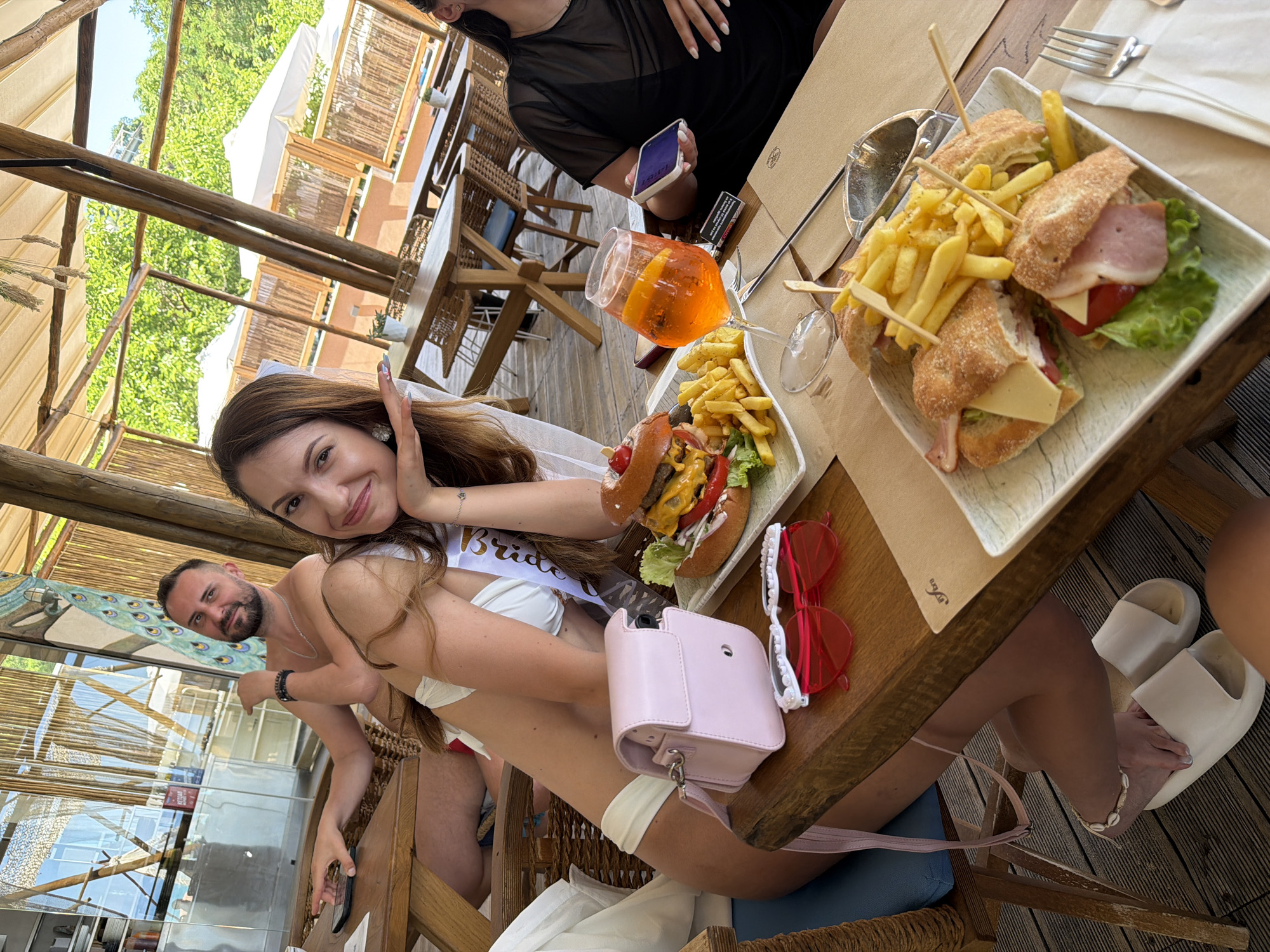 In the city of Kassandra Municipal Unit, Greece, a group of people is dining at a beachfront restaurant. In the foreground, a woman in a white bikini is seated at a table with a light pink purse and red heart-shaped sunglasses. In front of her, a hamburger and fries are served on a white plate, alongside a glass of orange beverage. To the right, another woman in a black top is holding a smartphone. Behind them, a man with a beard is seated, looking towards the camera. The restaurant features an outdoor seating area with wooden tables and chairs, surrounded by lush greenery and white umbrellas. The overall atmosphere suggests a casual and relaxed dining experience by the sea.