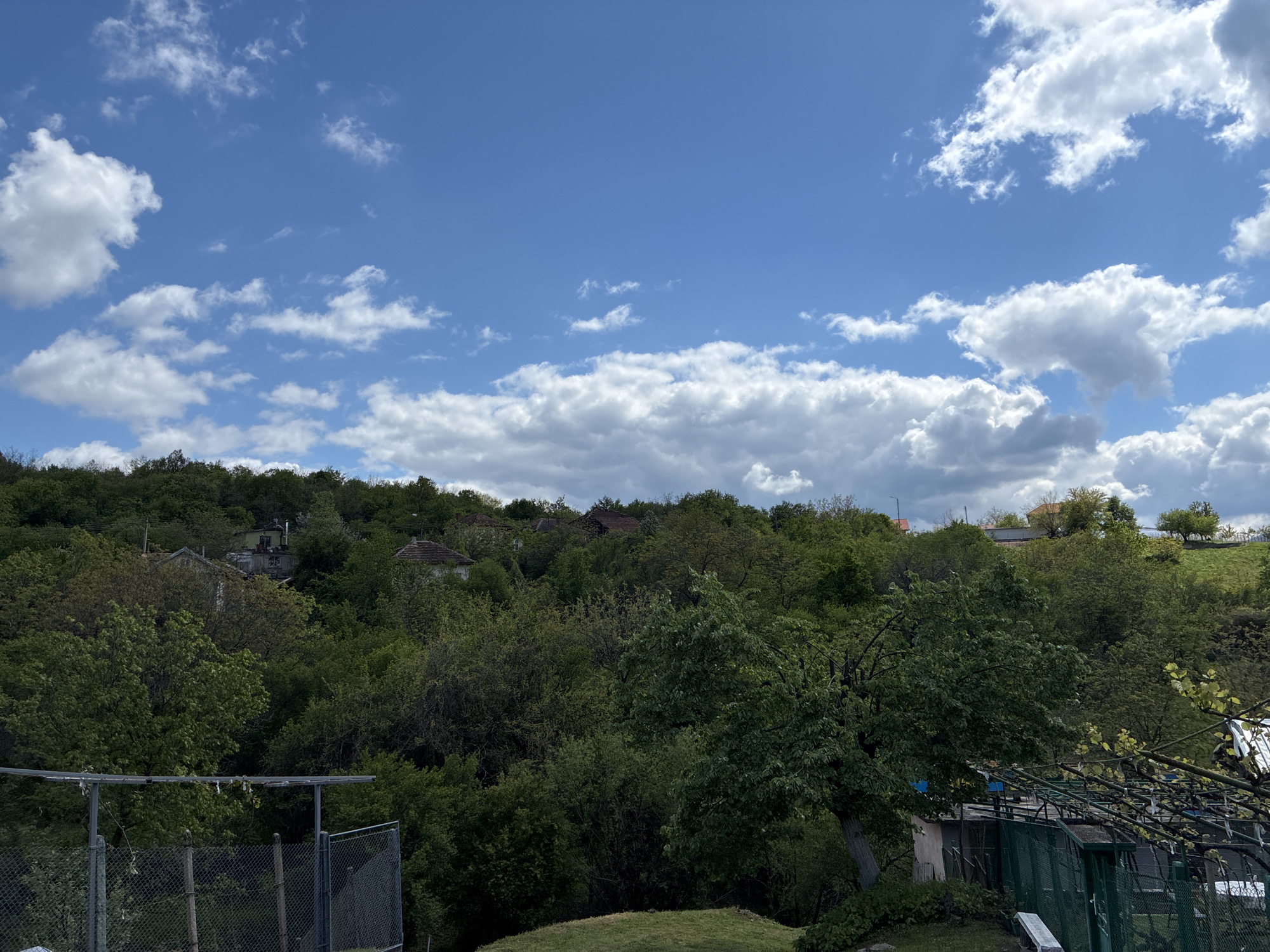 The image depicts a landscape in Bokilovtsi, Bulgaria, featuring a foreground with greenery, including trees and shrubs, and what appears to be a fenced-off area. Behind the immediate foreground is a densely vegetated hillside, interspersed with houses or buildings, partially obscured by trees. The sky is predominantly blue with scattered white clouds.