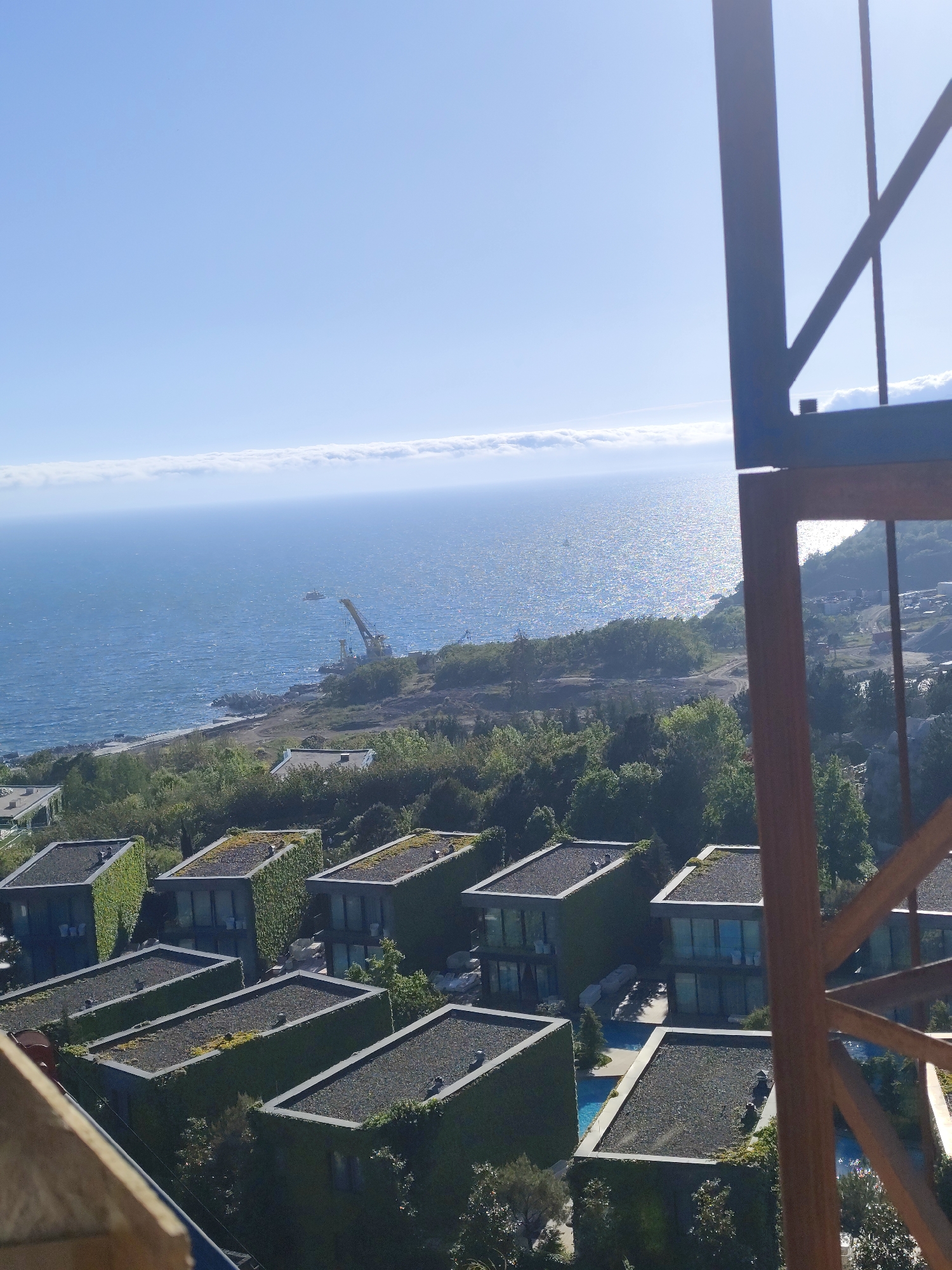 The image shows a coastal landscape with a series of modern, boxy houses, each appearing to have a flat roof covered with a dark material. The sides of the houses are heavily covered with green vines. A blue swimming pool is visible between the houses. Beyond the houses is a stretch of shoreline with vegetation, leading to a body of water, likely a sea or ocean, with a visible coastline in the distance. In the water, a crane on the shore can be identified. The sky is clear blue with some clouds along the horizon. A construction structure of some kind is visible in the foreground, partially obscuring the view. The location is Симеизский поселковый совет, Ukraine.