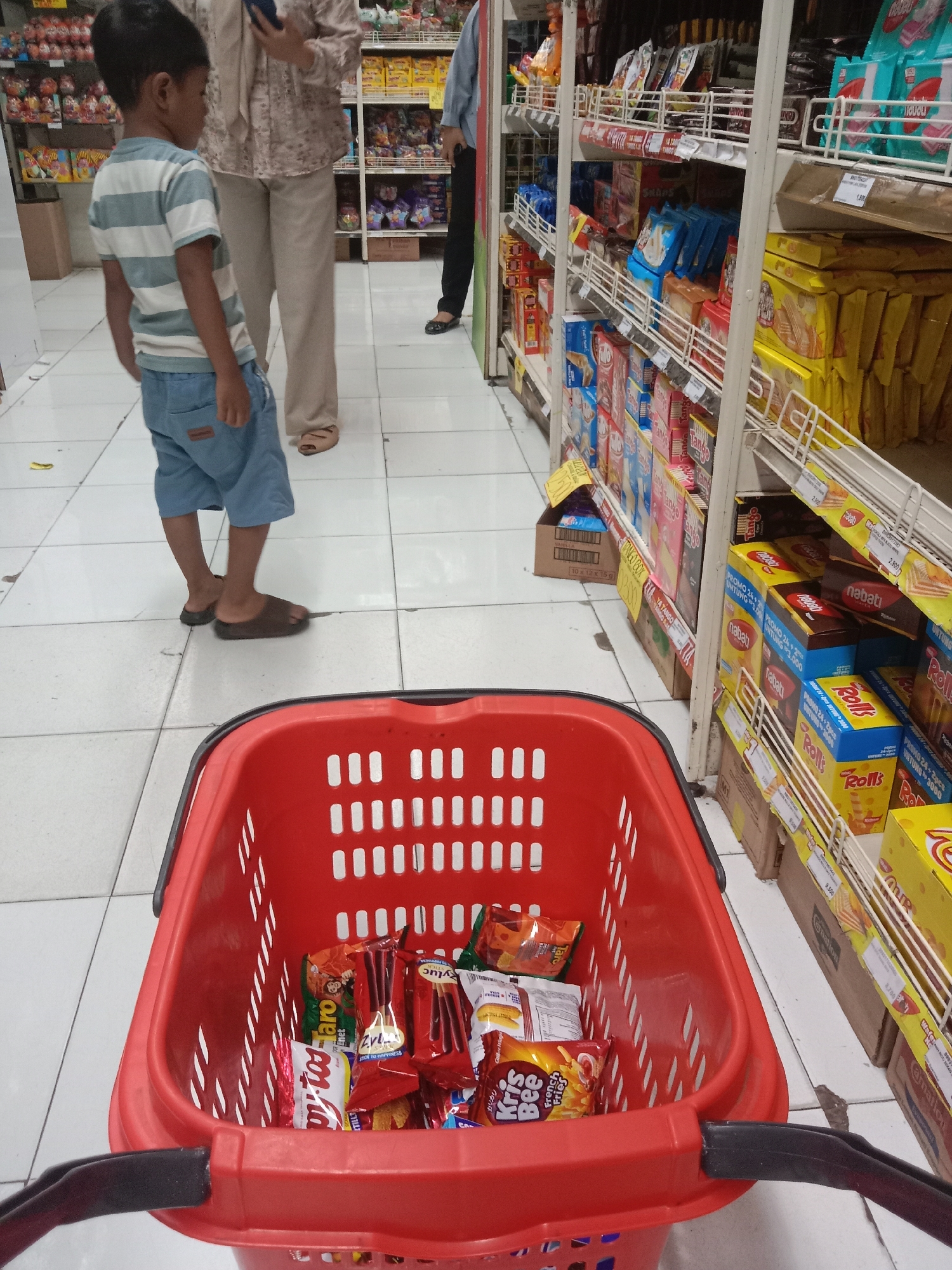 The image captures a scene inside a well-lit supermarket aisle, likely located in Probolinggo, Indonesia, as suggested by the surrounding product packaging that features Indonesian text. The perspective is from behind a large, red plastic shopping basket, which is filled with various snack food items. In the foreground, the basket's handles are visible.
The aisle displays numerous shelves stocked with packaged goods, primarily snacks like biscuits, crackers, and chips. Products from brands such as "Nabati," "Rolls," and "Krisbee" are identifiable. The shelves are constructed from white metal, and the flooring consists of white tiled squares.
In the mid-ground, a young boy, appearing to be around 5-7 years old, stands facing away from the camera. He is wearing a striped t-shirt with blue and white horizontal bands and blue denim shorts. He is barefoot or wearing very minimalist footwear. To his right, a woman, likely his mother or guardian, is partially visible, standing and looking down at her mobile phone. She is wearing a cream-colored patterned blouse and light-colored pants. Further back, another person is seen standing, partially obscured by shelves, wearing a light blue long-sleeved shirt and dark pants.
The overall atmosphere suggests a typical shopping trip, with the focus on the products within the basket and the shoppers in the aisle. The lighting is bright, characteristic of a retail environment.