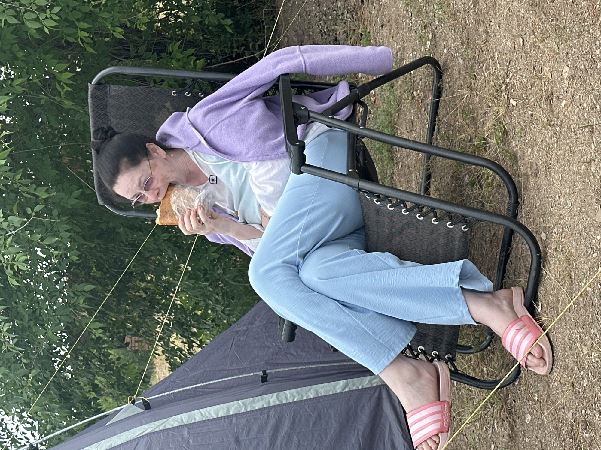 A woman is sitting in a reclining chair and eating a sandwich. She is wearing a light blue shirt, a purple hoodie, and light blue pants. She has pink slide sandals on her feet. The chair is black with a diamond-patterned fabric. A dark gray tent is visible to the left of the chair. The background consists of green trees and foliage. The ground is covered with dry grass and dirt. The scene appears to be an outdoor campsite.