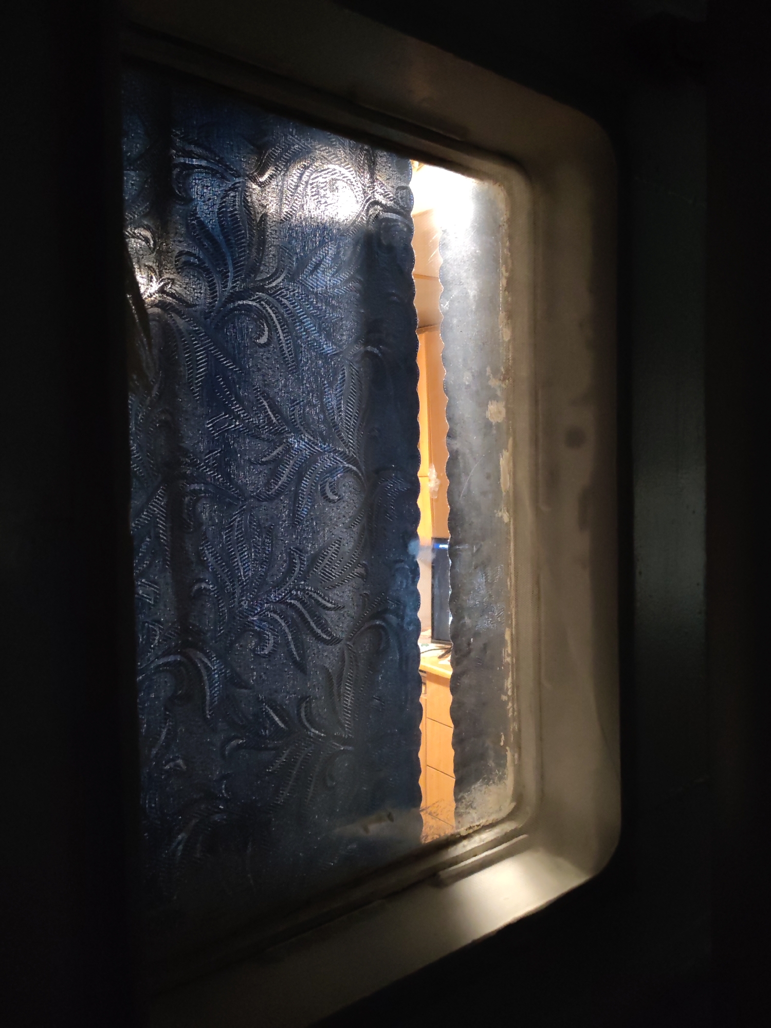 A close-up, low-light photograph shows a framed window with patterned glass. The glass obscures the view outside, but hints of an interior room are visible through a small gap. This room appears to have light-colored wood furniture and a television screen is visible in the background. The window frame is thick, painted in a light color. It shows signs of age and wear. The surrounding environment is dark, suggesting the photograph was taken in a dimly lit space, likely indoors. There's no information in the image to determine if the picture was taken in Tver, Russia.