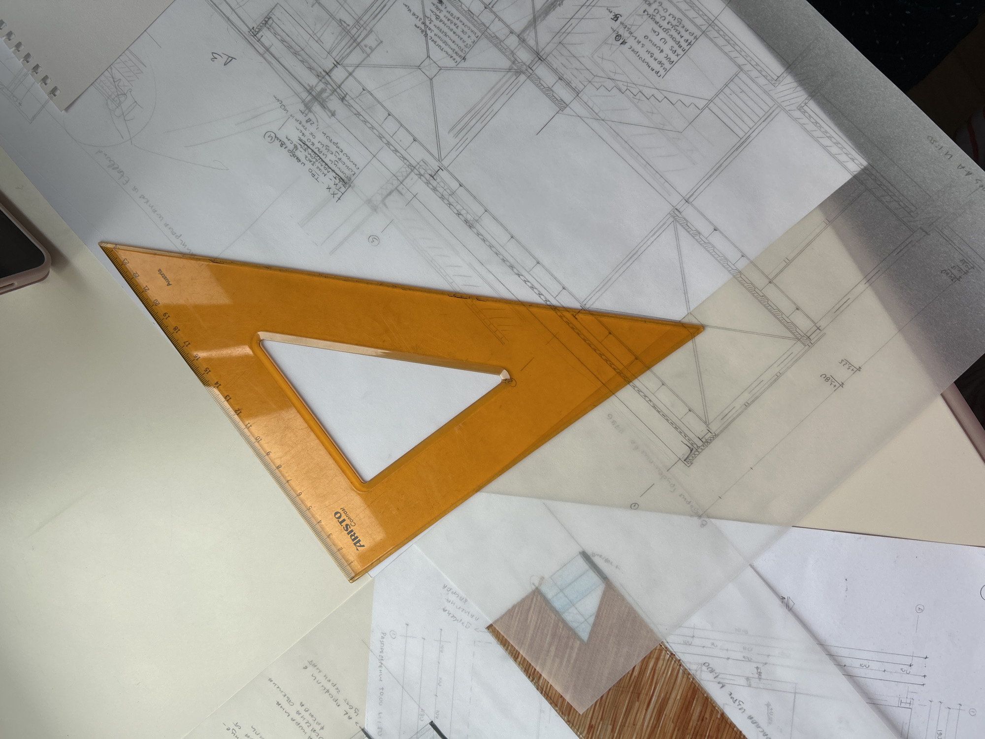The image shows an architectural design in progress. The focal point is a bright orange triangular ruler resting on several sheets of paper filled with architectural drawings and handwritten notes. The drawings appear to be technical blueprints or floor plans. Some of the notes are in Cyrillic. Without additional information, it's impossible to ascertain the location or the specifics of the project.