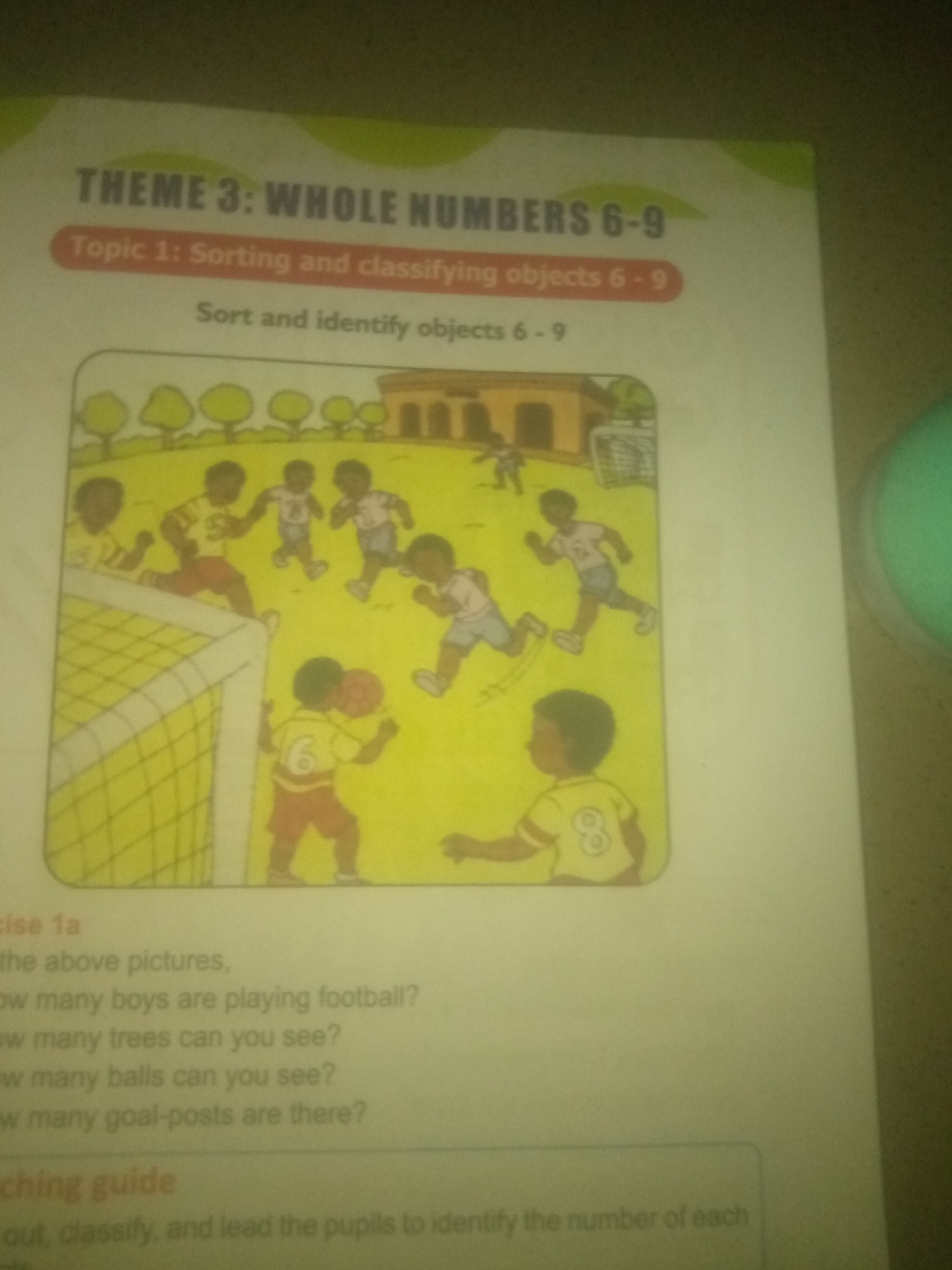 The image shows a page from a children's workbook. The title "THEME 3: WHOLE NUMBERS 6-9" is visible at the top, followed by "Topic 1: Sorting and classifying objects 6-9" and "Sort and identify objects 6-9". Below this is a colorful illustration of a group of children playing soccer on a grassy field. There are trees and a building in the background. The children are depicted in various poses, some running, some kicking a ball, and one child is near a soccer goal. The page also contains text related to an exercise, with questions such as "How many boys are playing football?", "How many trees can you see?", "How many balls can you see?", and "How many goal-posts are there?". Further text mentions "aching guide" and instructions for teachers. There is no information or imagery present that can be used to identify the location as Jauro Yinu, Nigeria, or any specific city.