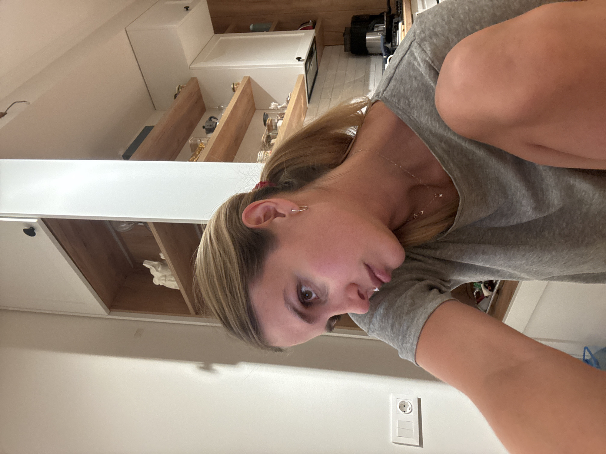 A woman is indoors in a kitchen. The kitchen has white cabinets, wooden shelves, and some appliances. The woman has blonde hair and is wearing a gray shirt. There is an electrical outlet on the wall. The location is in the city of Unknown, Unknown.