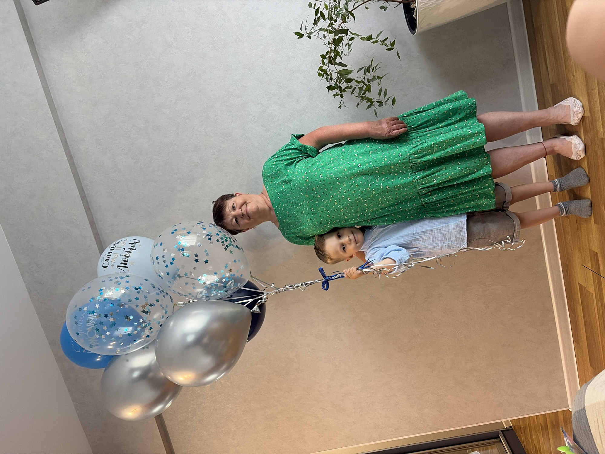 The image shows an elderly woman and a young boy holding a cluster of balloons. The woman, wearing a green floral dress, is smiling at the camera, while the boy, in a light blue shirt and grey shorts, looks towards the viewer. The balloons are a mix of silver, blue, and clear with stars, some featuring Cyrillic text. One balloon has the inscription "Сынок, мы тебя любим!" which translates to "Son, we love you!". The scene appears to be indoors, with a textured grey wall in the background and a wooden floor. A potted plant with green leaves is visible in the upper left corner. The lighting suggests it could be daytime. The overall mood seems celebratory and affectionate, possibly for a birthday or other special occasion.