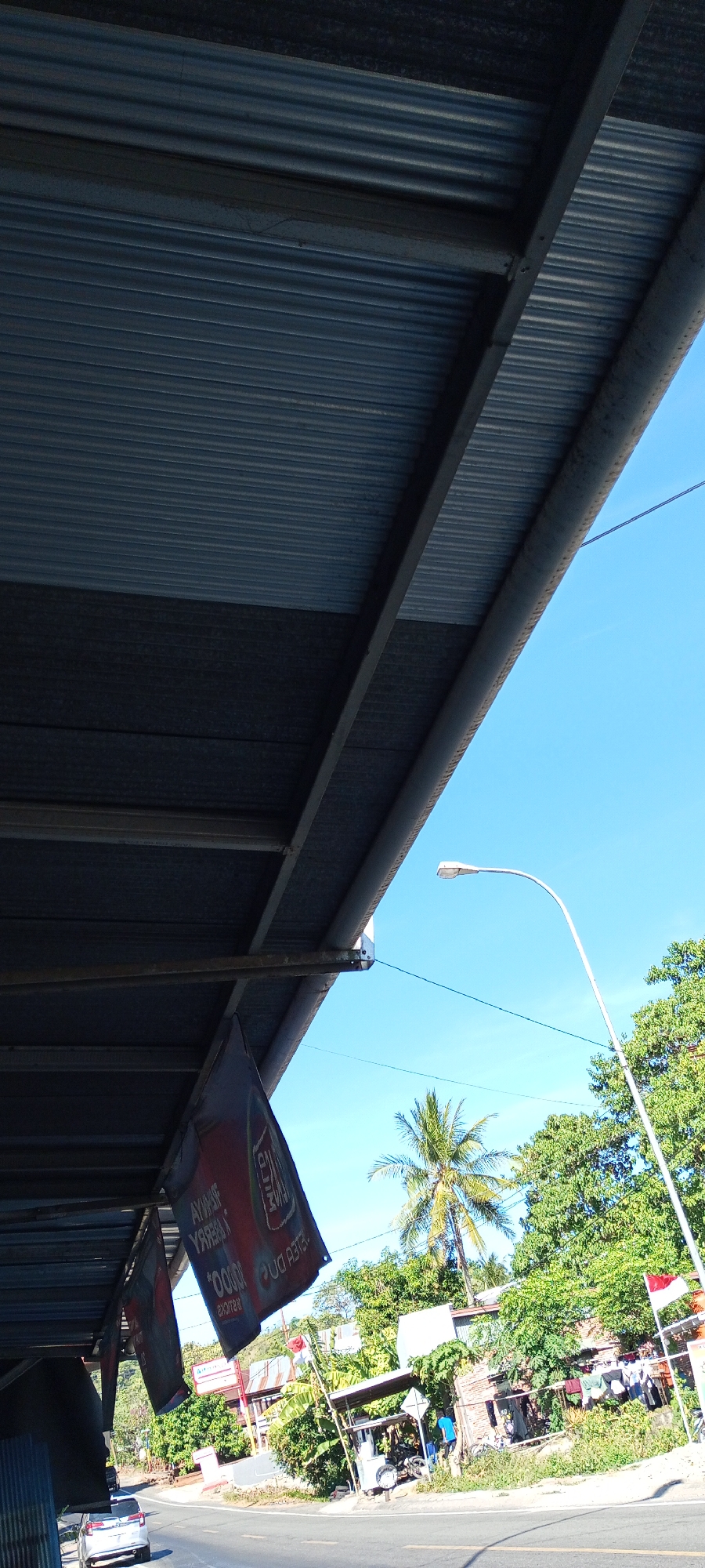 The image depicts a street scene in Pakkanna, Indonesia. In the foreground, a corrugated metal overhang shades a street. Two red and white flags are attached to the overhang, with text visible on one. A white car is parked on the left side of the road, and a motorcycle is parked on the right. A street lamp is visible against a clear blue sky. Lush green trees and buildings line the opposite side of the road. The scene is captured on a sunny day.