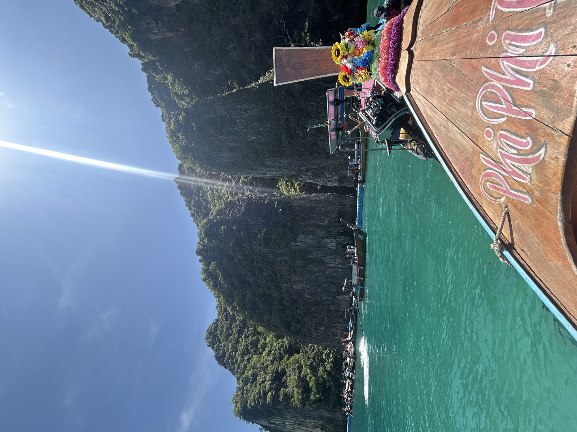 This is a medium shot, taken from the perspective of a boat, looking towards towering limestone cliffs and turquoise water. The boat in the foreground is wooden with "Phi Phi" painted in pink cursive on its bow. A colorful lei of flowers is draped over the engine of the boat. In the middle ground, numerous other long-tail boats are gathered in the clear, green water, some with passengers visible. A blue barrier or floating dock is visible in the water, separating some of the boats. The cliffs are covered in lush green vegetation and appear to be part of a bay or inlet. The sky is a bright blue with a single, strong sunbeam cutting diagonally across the upper right portion of the image. The overall scene depicts a popular tourist destination in Ao Nang, Thailand, likely a cove or lagoon accessible by boat.