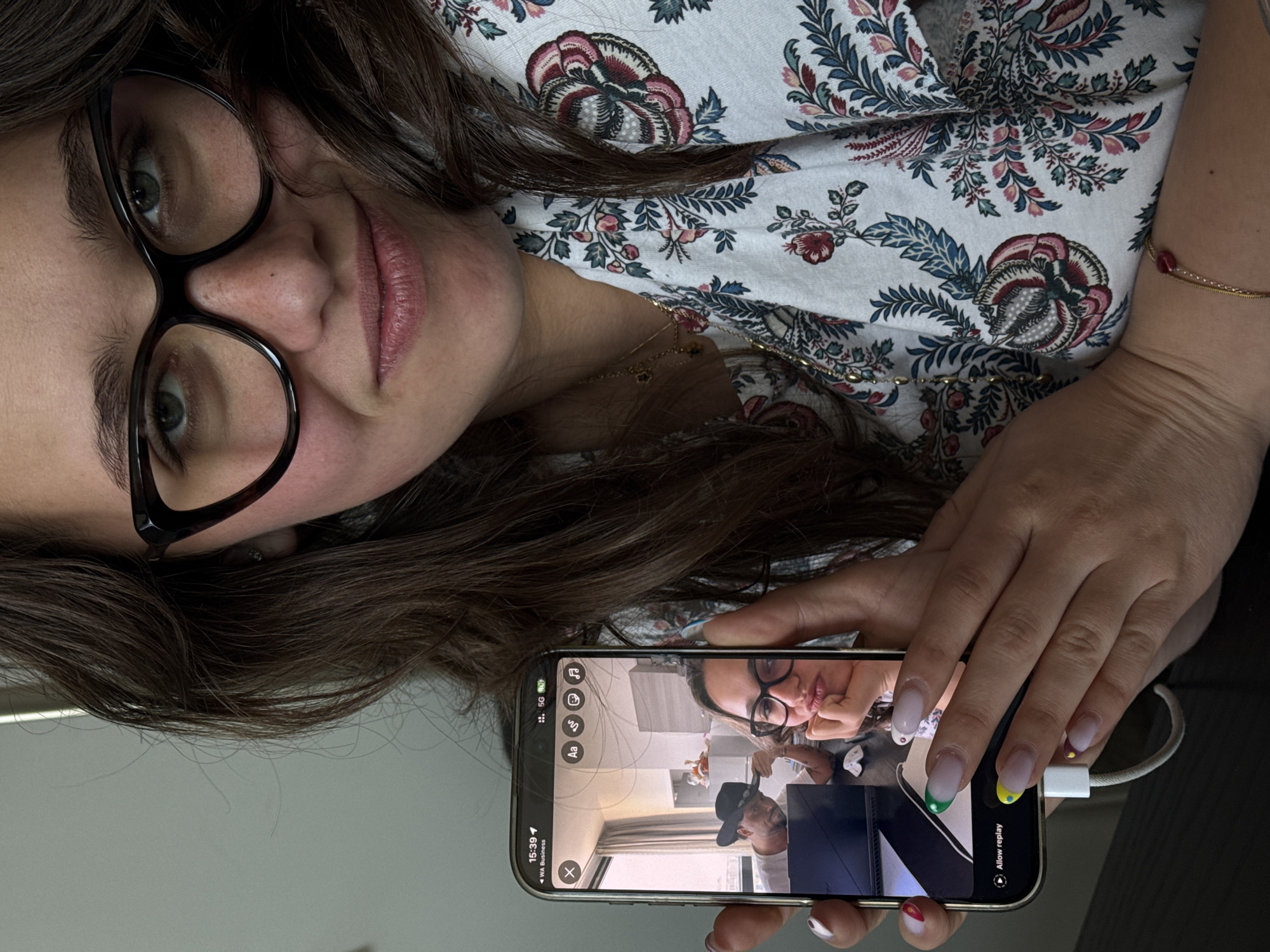A woman is depicted in a close-up, top-down shot, holding a smartphone that displays a video call or recording. Her face is turned slightly to the right, with dark brown hair framing her face and shoulders. She wears black-rimmed glasses, and her eyes are directed towards the phone screen or the viewer. Her lips are slightly parted, revealing a natural lip color. She is dressed in a light-colored top adorned with a detailed floral and paisley pattern in shades of blue, red, and cream. A delicate gold chain necklace with small dark beads and a matching bracelet with a red bead are visible.
Her hands, with French-manicured nails featuring colorful tips (green, yellow, and red/white), firmly hold the smartphone. A white charging cable is plugged into the phone. The phone screen shows a video, where a man wearing a black hat and a light-colored shirt is visible in the foreground. In the background of the video, a room with light walls and some shelves can be seen. The woman's own reflection, wearing her glasses and floral top, is also visible on the phone screen, creating an "image within an image" effect.
The phone's interface displays the time "15:39", "WA Business" (likely WhatsApp Business), a 5G network indicator, and icons suggesting media and editing options, including "Allow replay". The background behind the woman is a plain, light grey surface. The scene takes place indoors, likely in Milan, Italy, depicting a moment of digital interaction or content review in the afternoon.