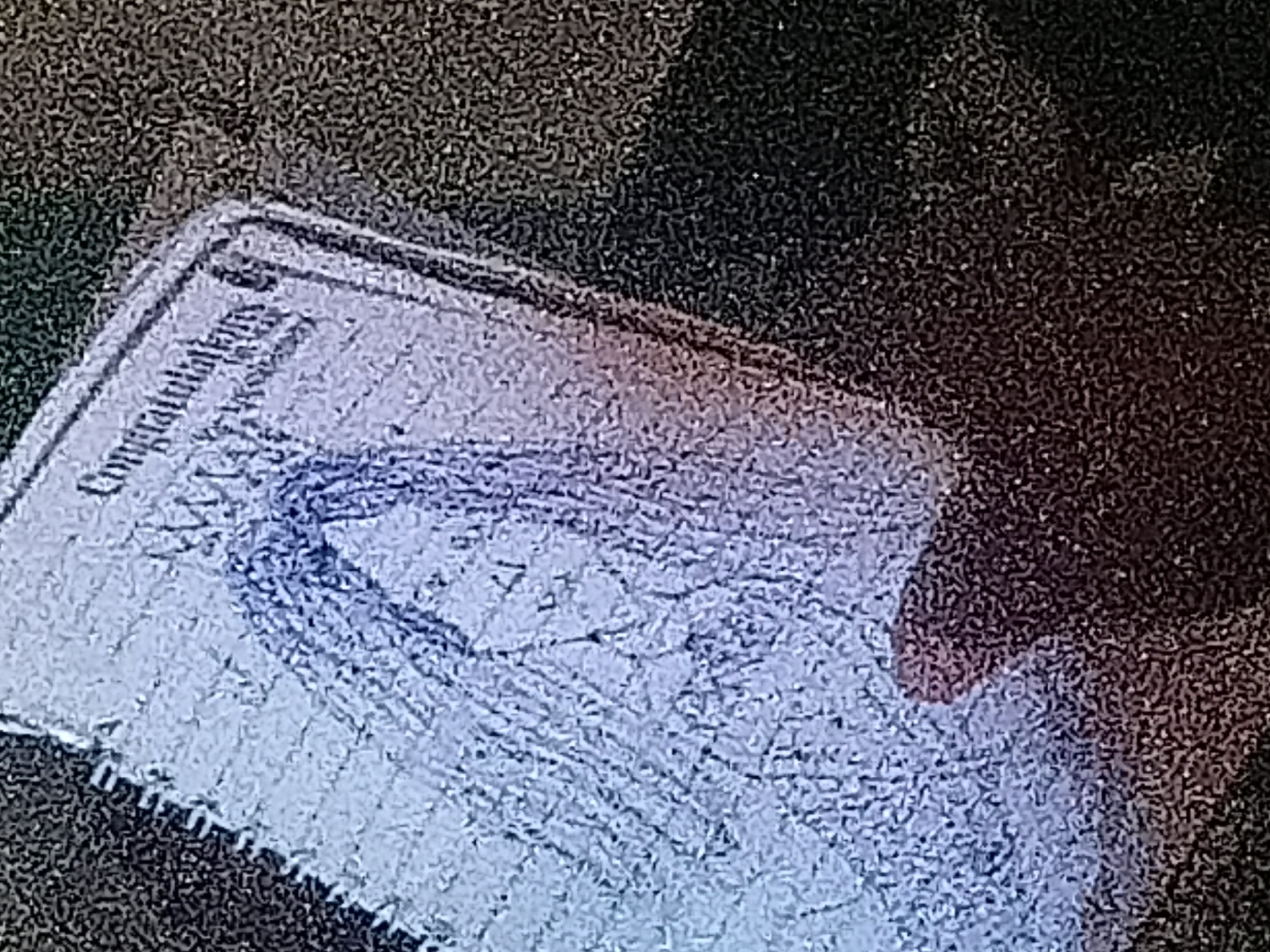 A low-resolution, grainy, close-up image depicts a light-colored rectangular paper or card held at a slight angle, tilted upwards to the right. The paper features the printed word "Congratulations" at its upper right corner. Below this text, a simple, hand-drawn sketch in blue ink or pencil shows a stylized figure, primarily characterized by long, flowing hair framing an indistinct face, with suggestions of a neck and shoulders. A dark shadow, consistent with a human hand, obscures the lower portion of the paper, indicating it is being held. The background is uniformly dark and features a coarse, pebbled texture. No identifiable markers or contextual elements within the image provide information regarding its specific geographic location, such as the city of Jalingo in Nigeria.