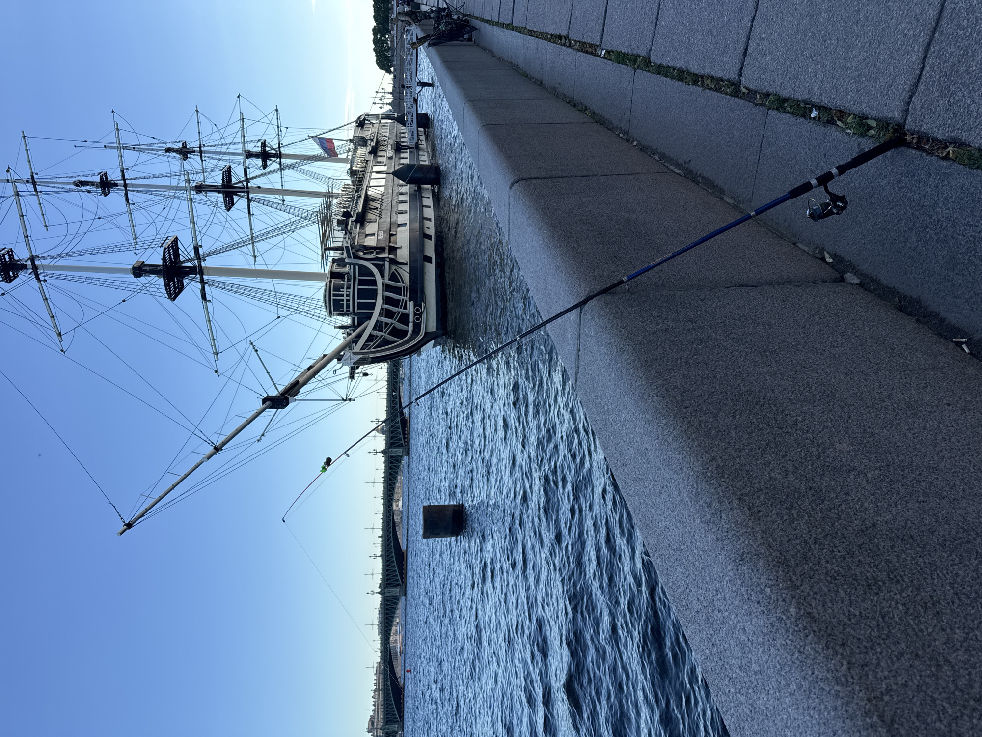 The image depicts a picturesque scene along the Neva River in Saint Petersburg, Russia. In the foreground, a fishing rod with a reel is propped against a grey granite embankment. The rod is dark blue with black and silver accents. Beyond the embankment, the Neva River flows with moderate ripples under a clear blue sky.
Dominating the middle ground is a large, historic-looking sailing ship docked on the river. The ship is multi-masted and appears to be a replica of an old vessel, with intricate rigging and wooden detailing. A Russian flag is visible on the ship's stern. In the distance, a long, green bridge spans the river, with buildings lining the far bank. A single, dark cylindrical buoy stands in the water to the left of the ship.
The scene is one of tranquil urban riverside activity, with the juxtaposition of the modern fishing equipment and the historical ship creating a sense of timelessness. The lighting suggests it is late afternoon or early evening, with the sun casting a soft glow on the water and the ship. There are no people clearly visible in the image, contributing to the serene atmosphere. The overall impression is one of a quiet moment by the water, with the grandeur of Saint Petersburg's architecture and maritime heritage serving as a backdrop.