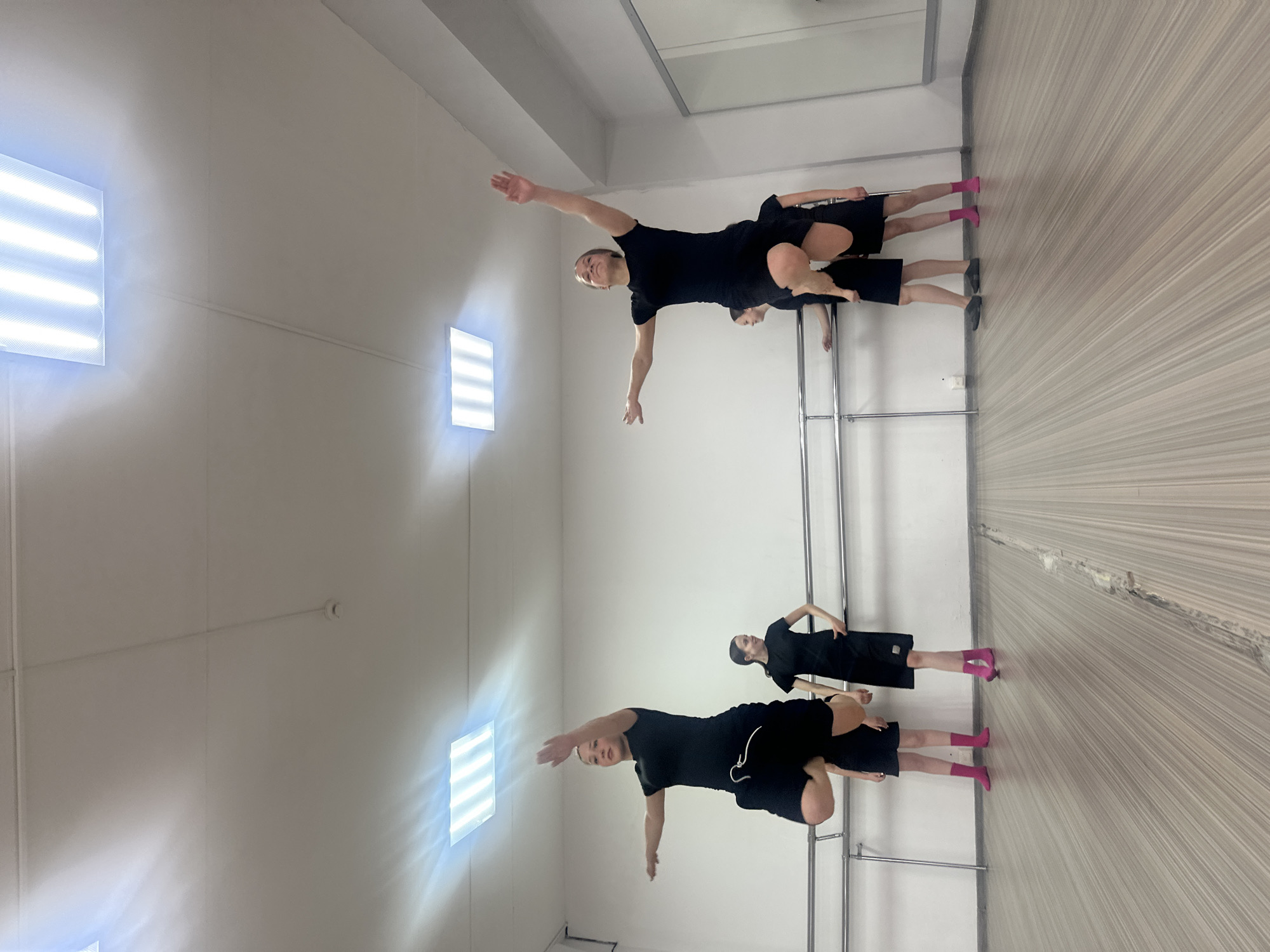 This image depicts an indoor scene, likely a dance or practice studio, located in Votkinsk, Russia. Four individuals, all dressed in black athletic wear consisting of t-shirts and shorts, are visible within the brightly lit space.
In the foreground, two individuals are actively performing a balanced pose, akin to an "airplane" or "superman" stance, with their arms extended horizontally and balancing on one leg. One of these individuals, towards the left, is wearing dark shoes or socks, while the other, towards the right, sports bright pink socks. Their facial expressions convey concentration.
The studio features a long, silver metal ballet barre installed along the right wall. This wall is also covered by a large mirror, which reflects the room and two additional people. One reflected figure is standing near the barre with hands on their hips, and another is partially visible, indicating a broader class or practice.
The room has a light-colored, possibly wooden or laminate, floor and plain, light-toned walls and ceiling. Two prominent, rectangular fluorescent light fixtures on the left wall provide ample illumination, creating an evenly lit environment for the activity. The setup strongly suggests a dedicated space for dance or physical training. There is no visible text in the image.