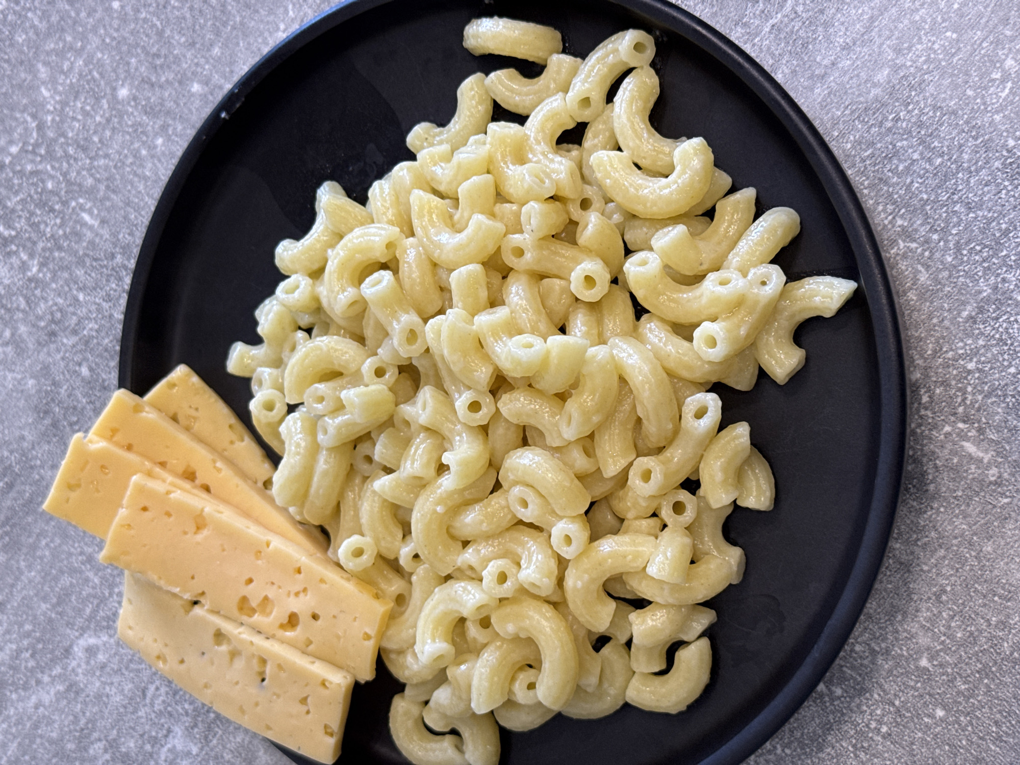 A close-up, overhead shot shows a black plate of elbow macaroni with cheese sauce. To the left of the pasta, on the plate, are four slices of light yellow cheese. The plate is set on a speckled gray countertop. The image does not provide any information about the city of Lyubertsy, Russia.
