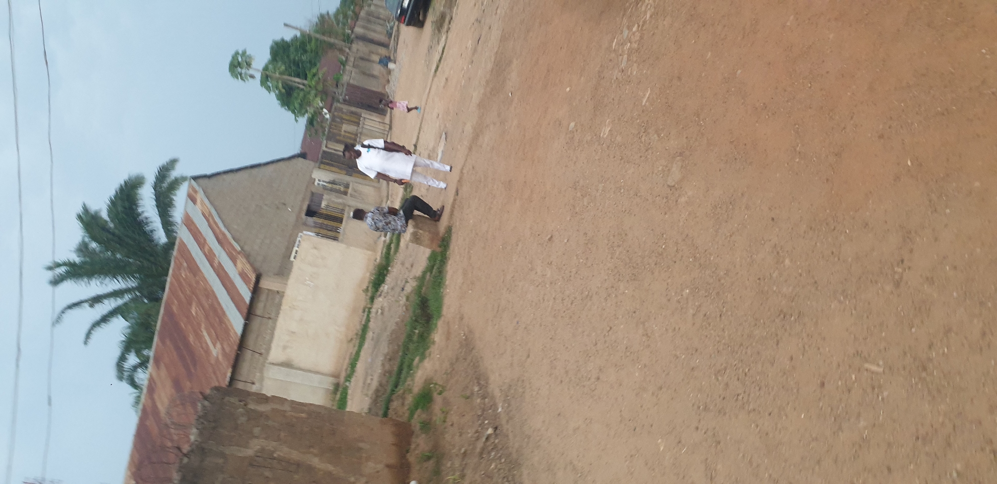 The image depicts an outdoor scene on a dirt road in Sabon Tasha, Nigeria. Two men are present, with one seated on a concrete block and the other standing. The seated man wears a patterned short-sleeved shirt and dark trousers. The standing man is dressed in a white traditional outfit. A young child in a pink dress is visible in the background, as is a dark-colored vehicle parked on the right side of the road. Structures with corrugated metal roofs and brick walls are present along the roadside, and vegetation, including palm trees, can be seen. The sky is overcast.