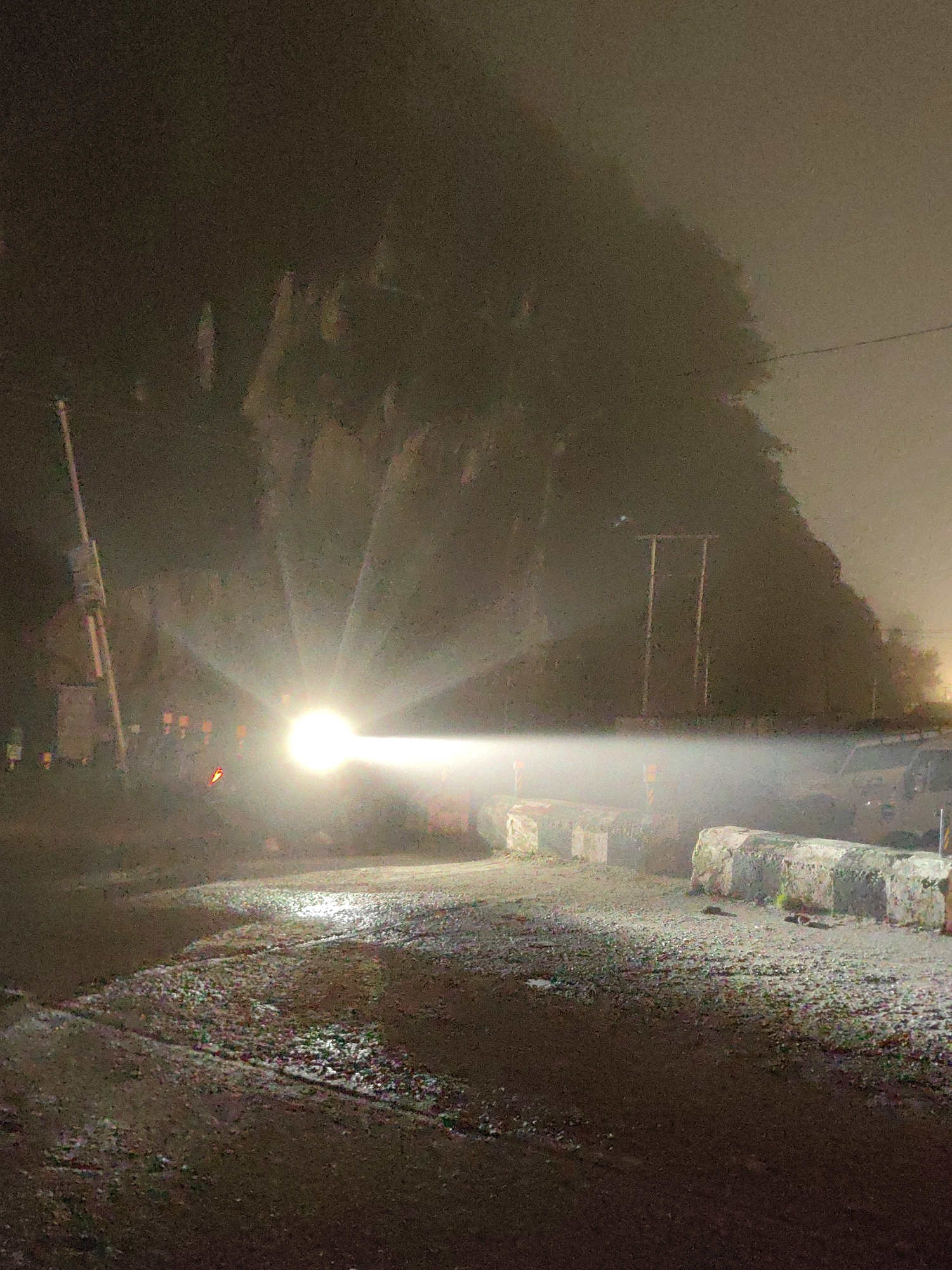 A bright headlight illuminates a foggy night scene on a road in Kulang, India. The light beam, emanating from an unseen vehicle, cuts through the thick mist, creating a hazy glow that obscures details in the distance. The road surface appears wet and reflective, with scattered debris and small stones. On the right side of the road, several large, concrete barriers are visible, some with reflective white and green stripes.
In the background, silhouetted trees and foliage form a dark, indistinct mass against the lighter, foggy sky. Two vertical poles, possibly part of a utility structure, stand out against the misty backdrop. On the left side of the image, a tall pole with signage is partially visible, along with a few distant, blurred figures. The overall atmosphere is eerie and atmospheric, suggesting a low visibility condition due to the fog. The time of day is clearly night, and the weather is foggy, contributing to the obscured visibility and sense of isolation. No text is discernible.