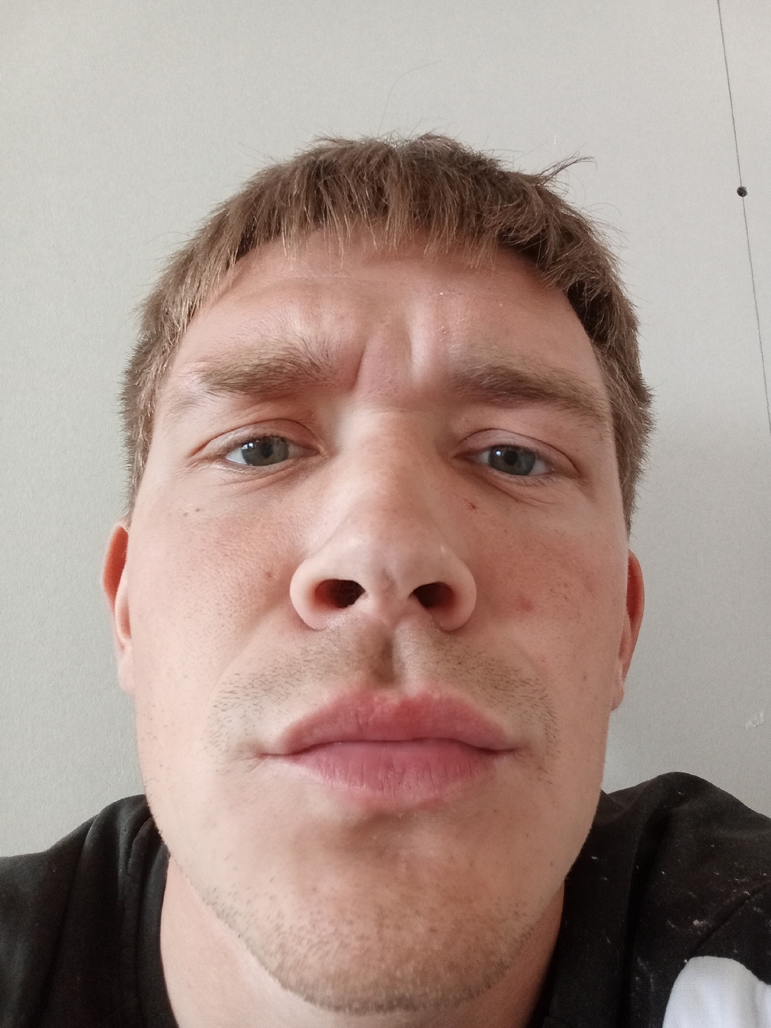 A close-up, low-angle selfie captures the face of a man, likely taken indoors in Chaikovsky, Russia. The man has short, light brown hair styled with a fringe, and distinctive blue eyes. His eyebrows are slightly furrowed, conveying a neutral to contemplative expression. He has light stubble visible on his upper lip and chin. His skin is fair, and his lips appear full. The man is wearing a dark t-shirt, with parts of it, including some white markings, visible at the bottom of the frame. The background is a plain, light-colored wall. In the upper right corner of the image, a thin wire or string hangs against the wall, featuring a small dark spherical object, possibly a bead or weight. The activity depicted is posing for a photograph. No text is visible in the image.