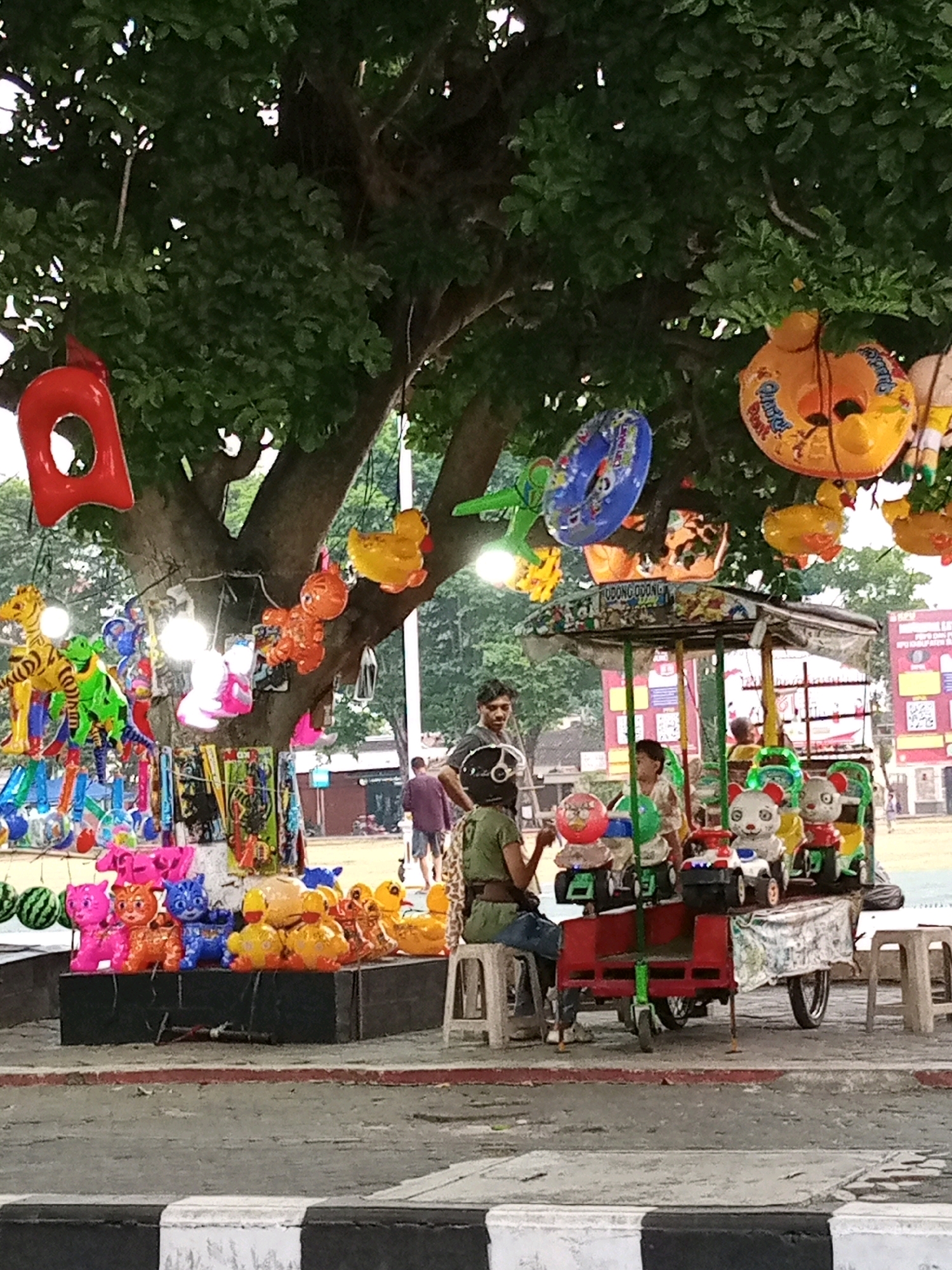 In Sukoharjo, Indonesia, a street vendor is selling inflatable toys and children's rideable toys from a cart under a large tree. A variety of colorful inflatable items, including ducks, cats, and rings, are hanging from the tree's branches. In front of the tree, a stand displays more inflatable toys, such as cats and ducks, arranged on a dark surface. The vendor's cart features several small, motorized rideable toys designed for children, some of which have animal-themed seats like bears. A few people are visible in the background, including a vendor operating the rideable toys and a child sitting on one of the toys. The scene appears to be a public outdoor space, possibly a park or a busy street.