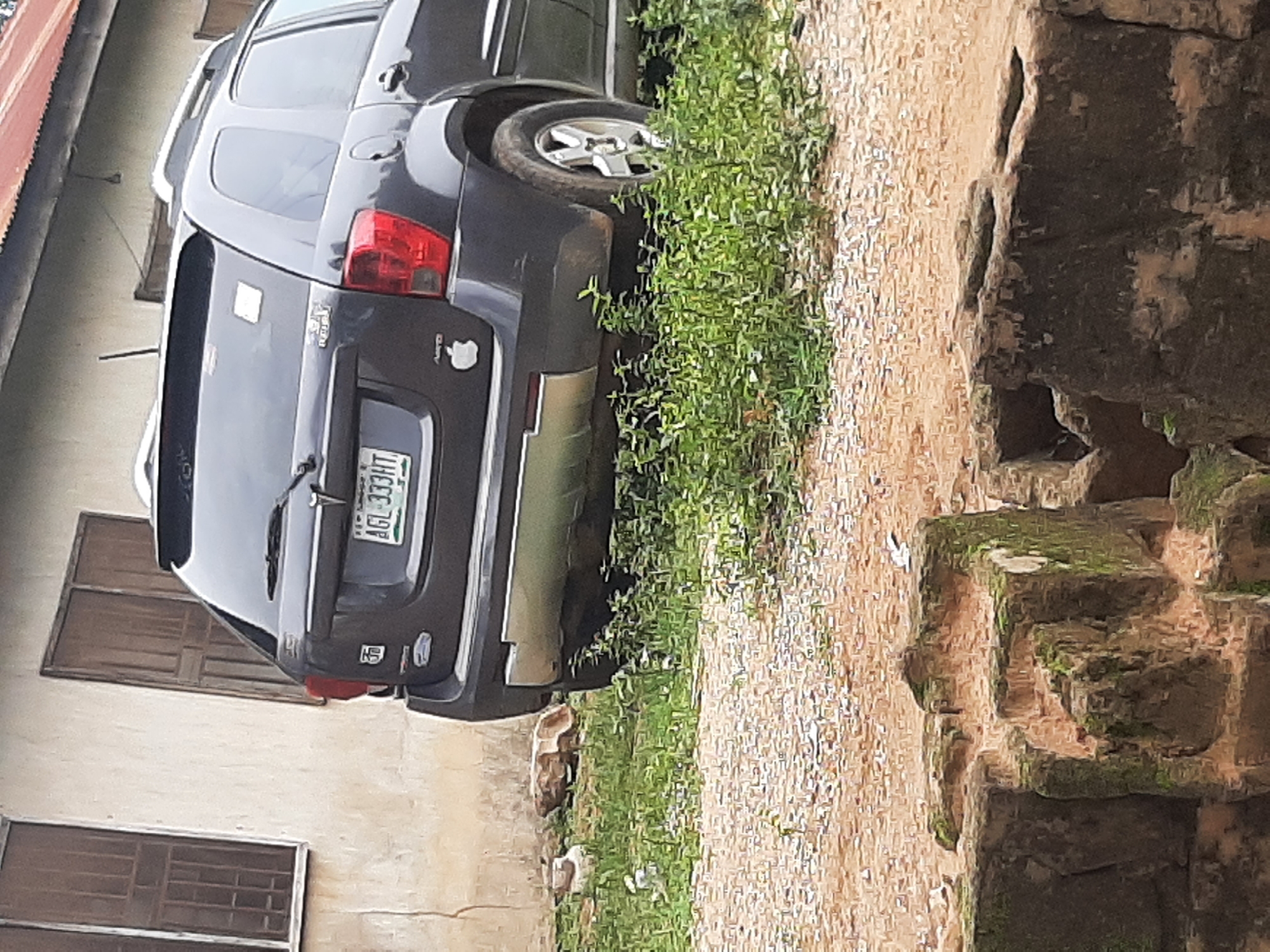 The image shows the rear of a dark gray SUV parked on a dirt and gravel surface with sparse green vegetation. The SUV has a license plate that reads "AGL-333HT". A white square sticker is visible on the rear hatch. To the left of the SUV, a building with light-colored walls and two brown-paned windows is visible. In the foreground, there are partially visible stone structures that appear weathered and moss-covered. The overall scene suggests an outdoor setting in a somewhat unkempt area. It is highly probable this image was taken in Ogbomosho, Nigeria, given the context provided.