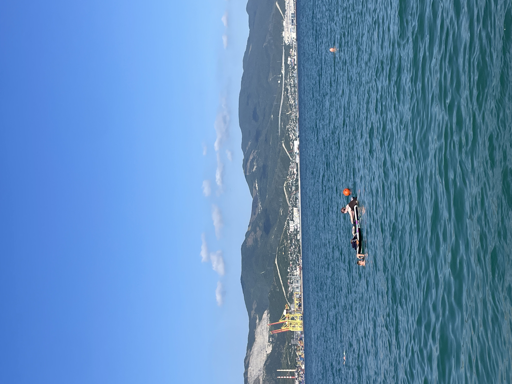 The image depicts a scenic coastal view in Sadovyi, Russia, under a clear blue sky with a few wispy clouds. The scene is divided into two main parts: a serene, deep blue ocean and a lush, mountainous coastline. The ocean is calm with gentle ripples, reflecting the bright daylight.
In the foreground of the ocean, a person is visible, seemingly participating in a water sport. They are lying on a small, dark-colored board with white accents, possibly a surfboard or a paddleboard, and appear to be wearing a dark wetsuit. A bright orange buoy floats near them, and another similar buoy is visible further in the distance.
The coastline features a densely populated area with buildings nestled against the base of a large, green mountain. A highway or road is visible snaking along the mountainside, indicating a well-developed area. In the lower part of the coastline, industrial structures, including tall cranes with yellow and red accents, are prominent, suggesting a port or industrial zone. The overall impression is one of a vibrant coastal town backed by dramatic natural scenery, with recreational activity taking place on the water. The time of day appears to be late morning or early afternoon, given the bright sunlight and clear skies, characteristic of fair weather.