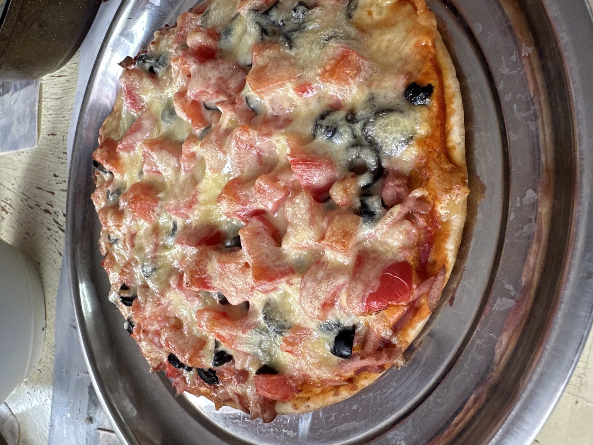 A round pizza with chopped tomatoes, black olives, and melted cheese is presented on a silver metal plate. The pizza has a thin crust and appears to be freshly baked. The plate is placed on a light-colored wooden surface. In the background, a white bowl and a dark pot are partially visible. The scene is indoors, likely in a domestic setting. There are no people visible in the image. The lighting suggests it is daytime. The image's location in Yaroslavka, Russia, is not discernible from the visual information provided.