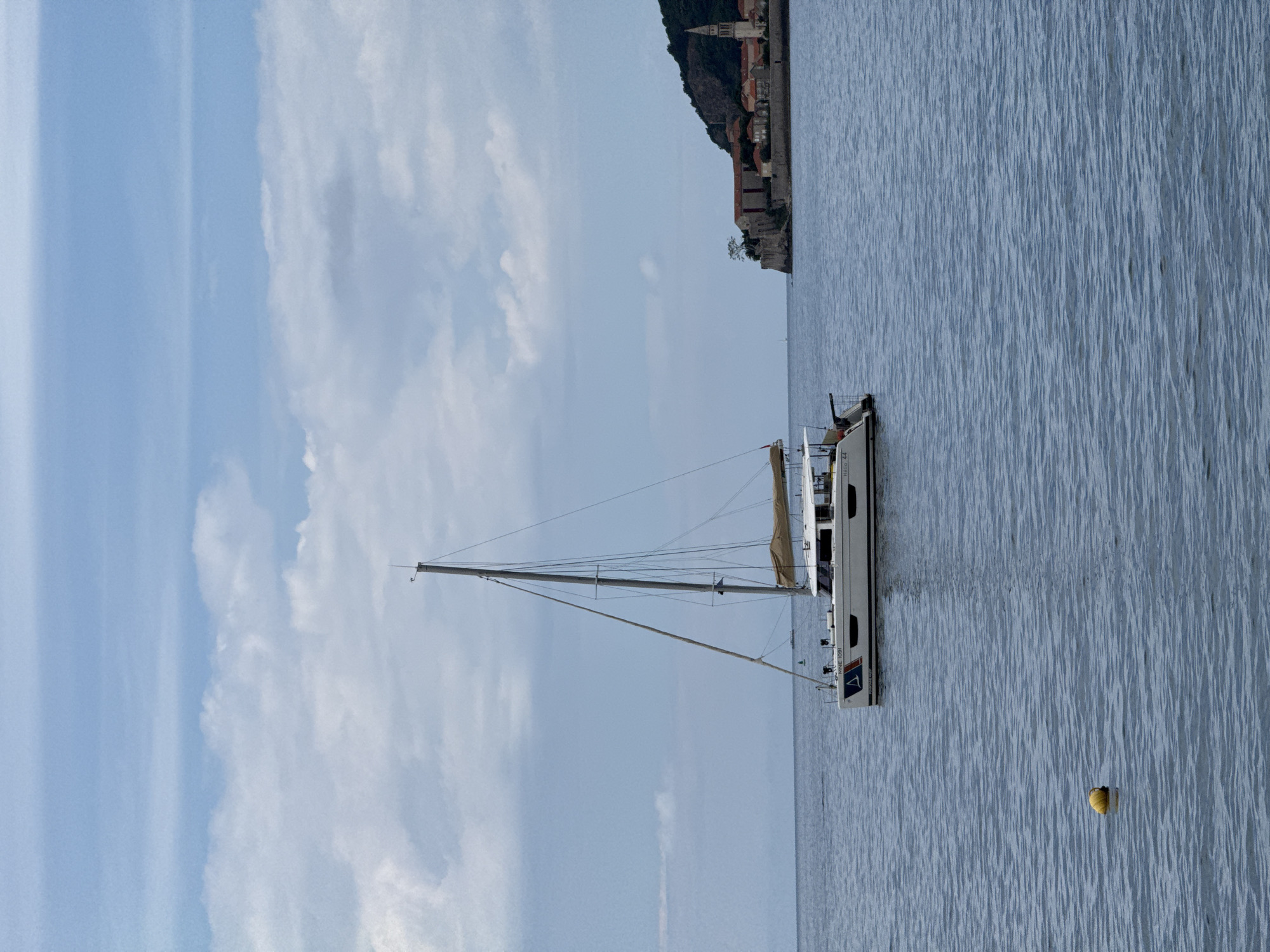A horizontal medium outdoor shot shows a white sailboat at anchor in the blue-gray waters of the Adriatic Sea near Budva, Montenegro.
The sailboat is positioned in the right half of the shot with its bow facing the top. The tan sail is unfurled. The sail bears the identification markings "Hello 44" and "NINE 1091." The mast and rigging reach up toward the upper-left. The boat casts a shadow on the water.
In the water to the right of the sailboat, a yellow buoy floats.
In the distance along the upper edge of the shot, the coastline of Budva, Montenegro, is visible, including a church spire atop a forested hill.
The sky above the sea is a light blue color with scattered white clouds.