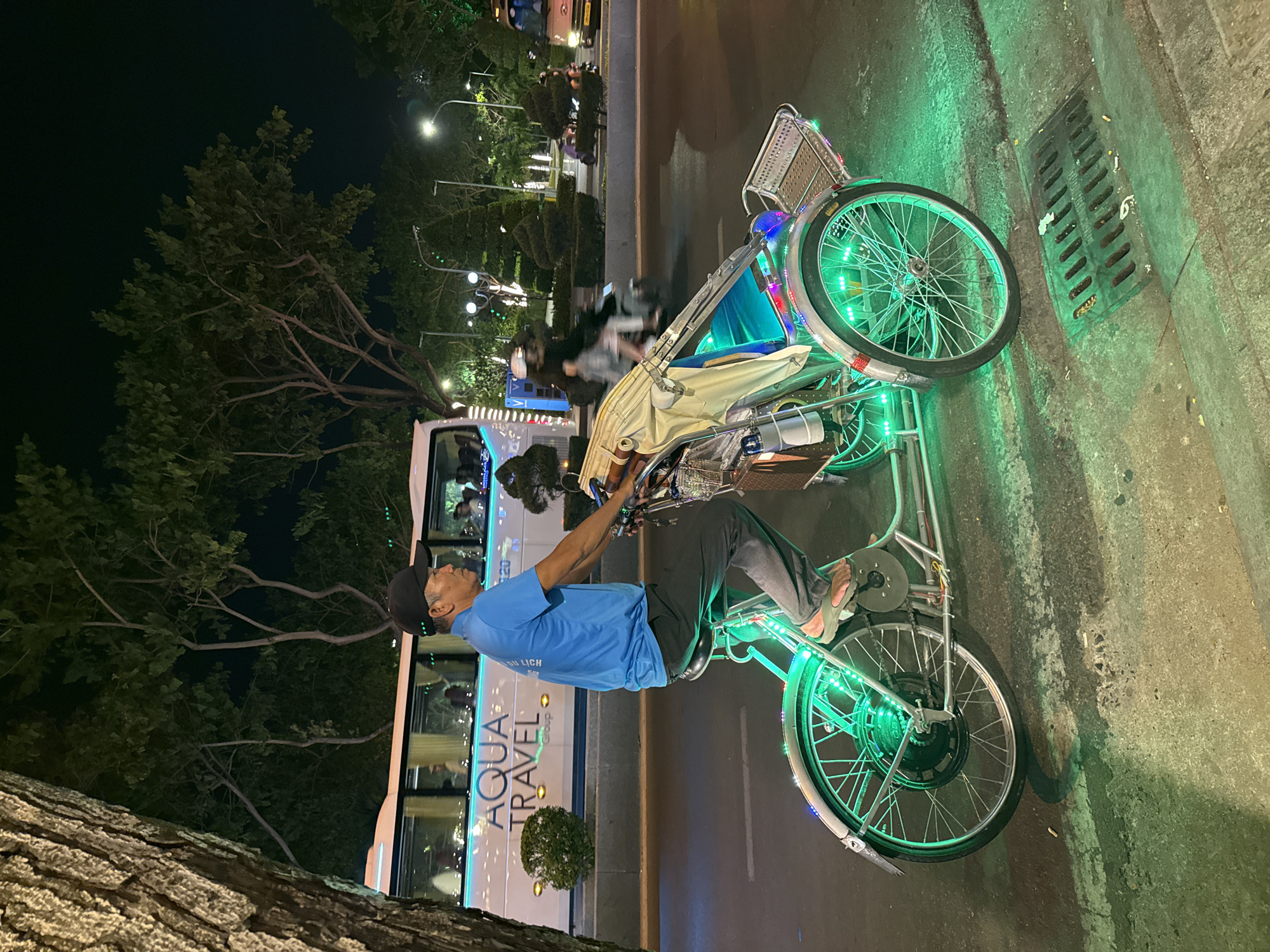 This media file depicts a night scene on an urban street, likely in Nha Trang, Vietnam. The main subject is a cyclo (pedicab) elaborately decorated with vibrant green LED lights on its wheels and frame, and additional colorful lights around its front passenger carriage, which is covered by a light-colored canopy. A wicker basket is attached to the front of the cyclo.
A man, presumably the driver, is seated on the cyclo. He wears a blue t-shirt, dark pants, and a dark baseball cap. His hands are near the handlebars. Text is visible on his shirt, appearing to be "DU LICH" (Vietnamese for tourism).
In the background, a large white tour bus is prominent, displaying "AQUA TRAVEL" and "Group" in illuminated text on its side. Several passengers are visible inside the bus. Further in the background, streetlights illuminate the street, where other faint figures and vehicles can be seen. Large trees with dark foliage line the left side of the street, and a textured tree trunk occupies the bottom-left foreground.
The cyclo appears to be stationary, positioned at the edge of the road next to a concrete sidewalk. A metal storm drain grate and some discarded cigarette butts are visible on the sidewalk near the cyclo's front wheel. The overall impression is a calm urban night scene, with the brightly lit cyclo suggesting it is either waiting for passengers or operating as part of a night tour experience. The time of day is clearly night due to the dark sky and prominent artificial lighting.