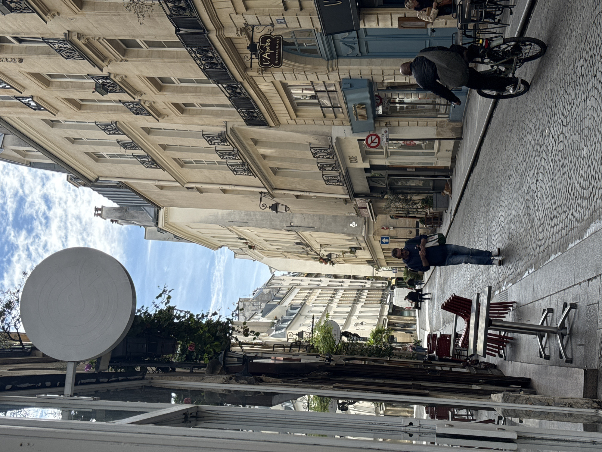 The image depicts a street in Paris, France. The street is lined with tall, light-colored buildings with ornate iron balconies. Cobblestones pave the street. A man is walking down the street, carrying something in his hands. A person on a bicycle is also present. Tables and chairs are set up outside a café or restaurant. A large, round sign is visible above a storefront. The sky is blue with some clouds.