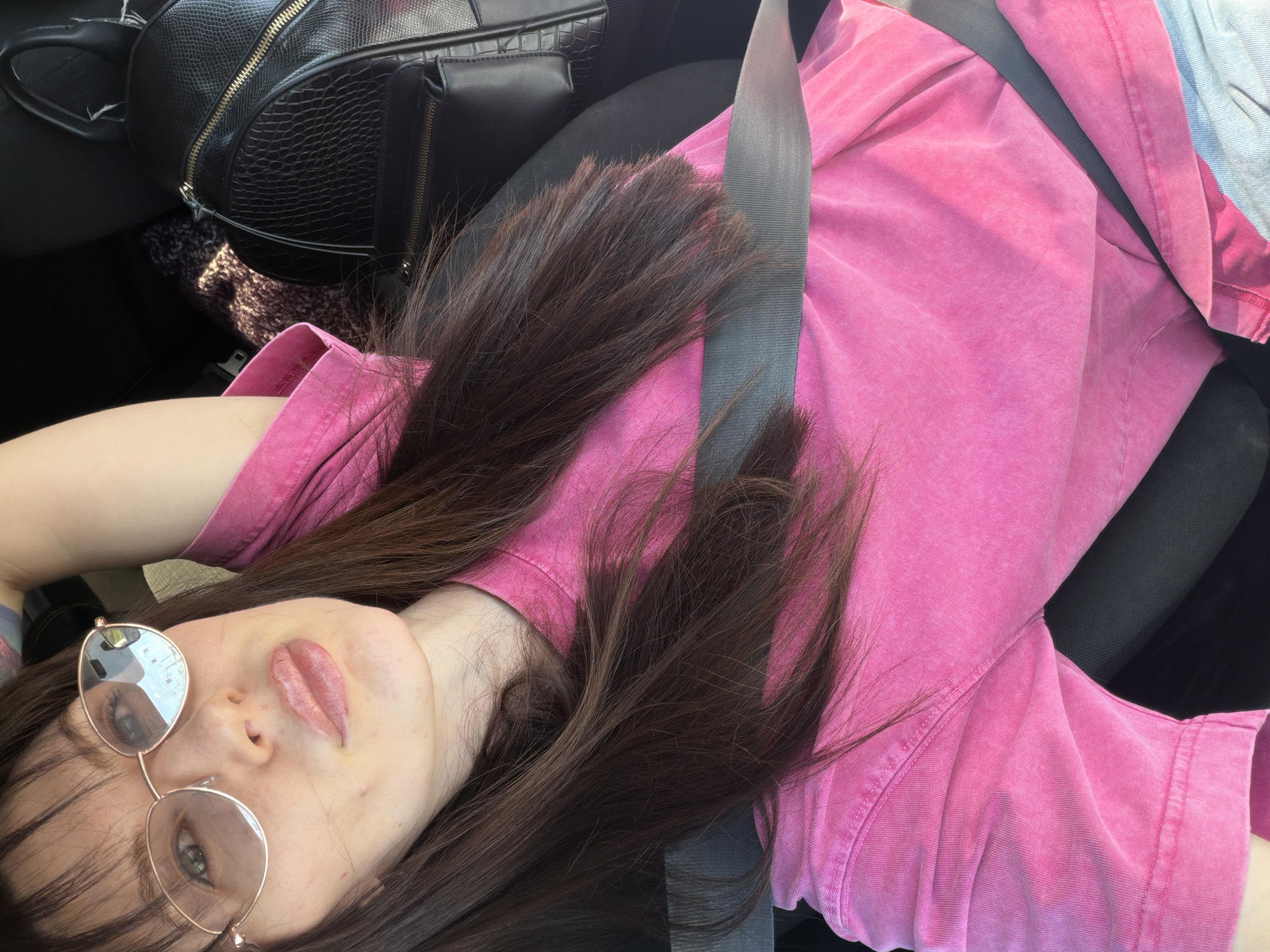 A young woman with long brown hair is reclined in a car seat, wearing a pink shirt and a seatbelt. She is wearing round glasses and appears to be looking at the camera. A black handbag is visible in the upper left corner of the frame. The location is inside a vehicle and it is not possible to determine if the city is Perm, Russia.